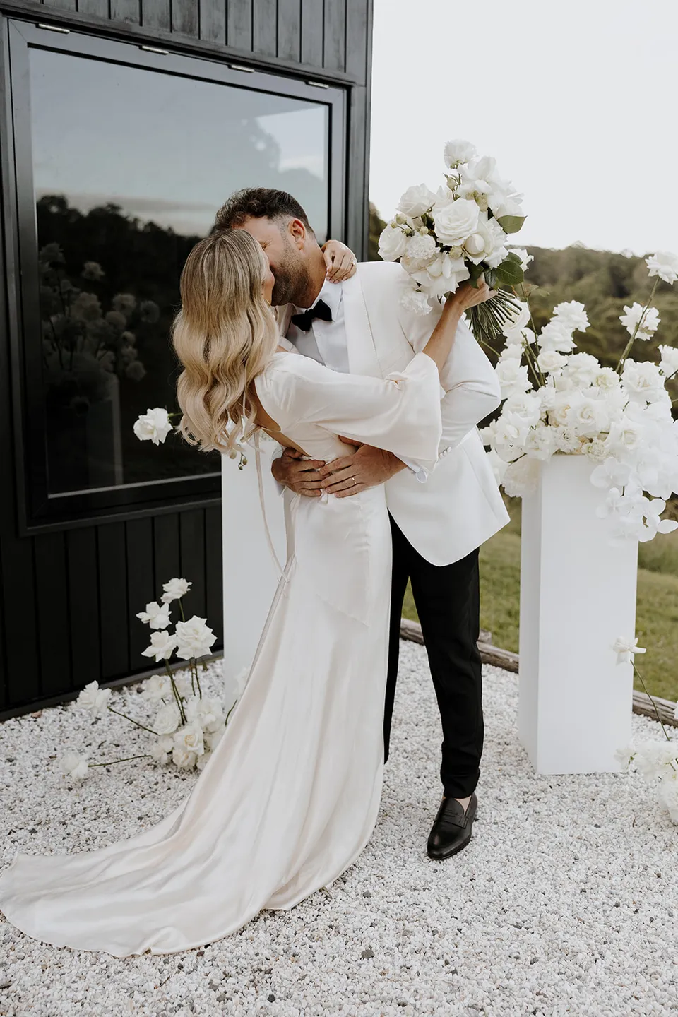 Greg embracing Gabrielle as they kiss beside the ceremony flowers, her white bouquet raised in her hand at Roundhill Retreat in Obi Obi, Sunshine Coast Hinterland, for their Elopement Collective elopement.