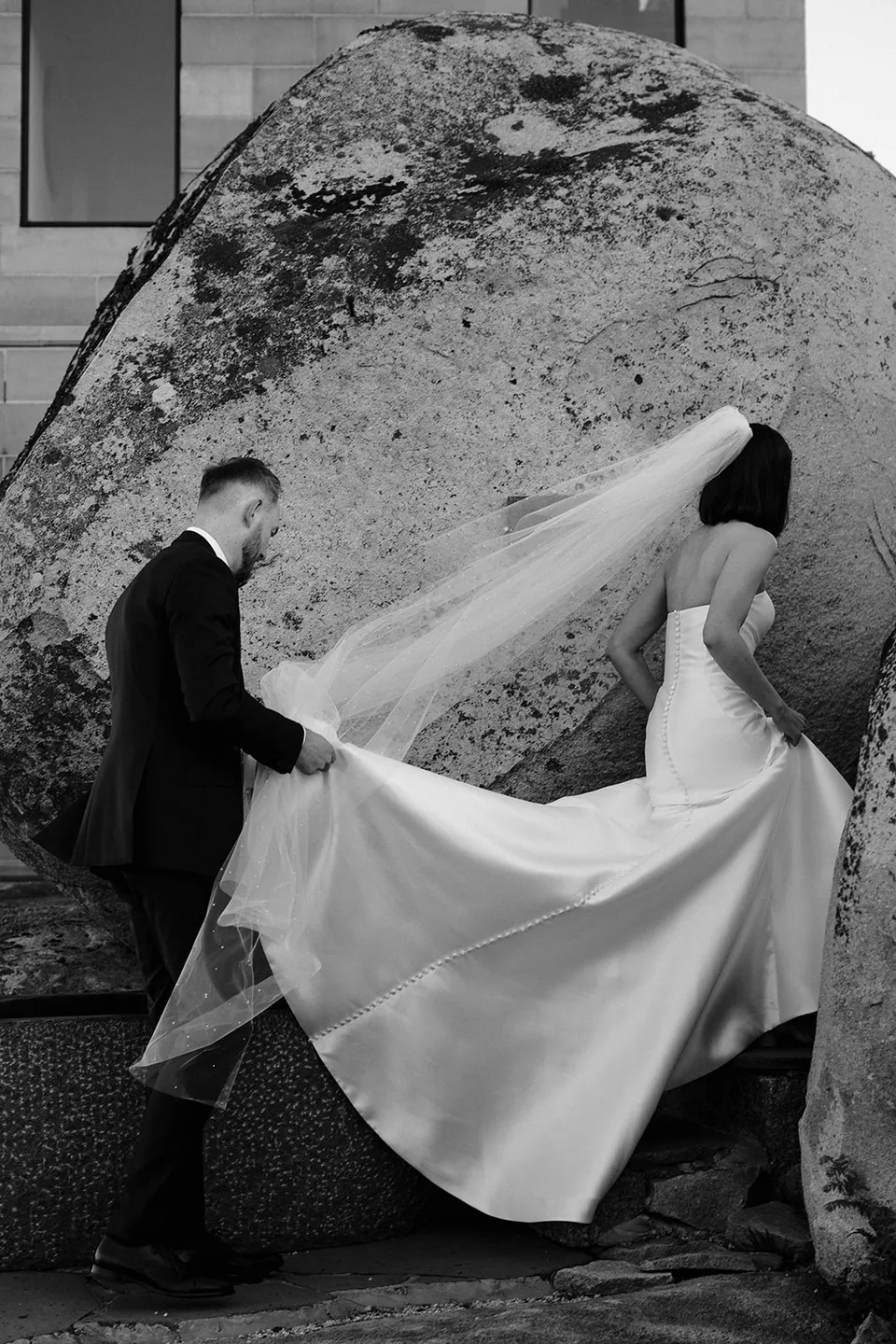 Black and white photograph of Ryan carrying Meg’s dress train as she makes her way between large rocks. At The Keep on Tasmania’s east coast, for an Elopement Collective elopement. Photograph by Jenna Agius.