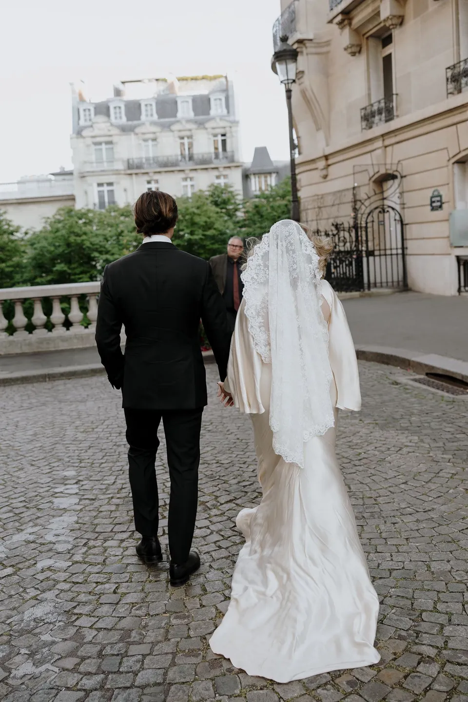 Rear view of Kara and Samuel holding hands as they walk towards celebrant Josh Withers, beside Ivy Bleu’s red floral ceremony setup, at Avenue de Camoëns in Paris, France, during an Elopement Collective elopement. Photograph by George Bowden.