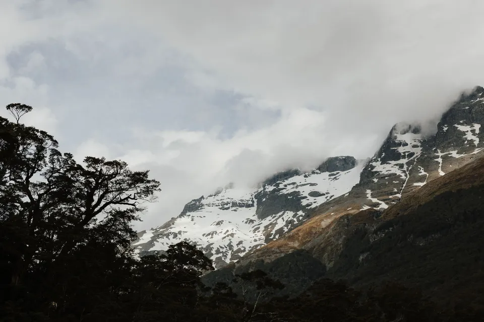 Snow-streaked mountain peaks rising above native forest beneath low clouds beside the ceremony field, in the field on the road to Paradise near Glenorchy, in the Queenstown region of New Zealand, for an Elopement Collective Queenstown elopement. Photograph by Bulb Creative.
