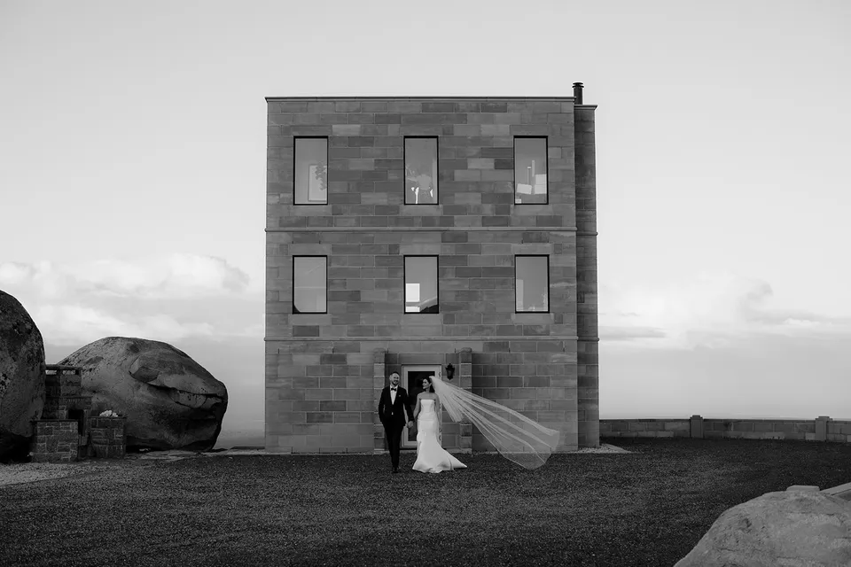 Black and white wide view of Meg and Ryan in front of The Keep’s sandstone tower, with her veil blown sideways. At The Keep on Tasmania’s east coast, for an Elopement Collective elopement. Photograph by Jenna Agius.
