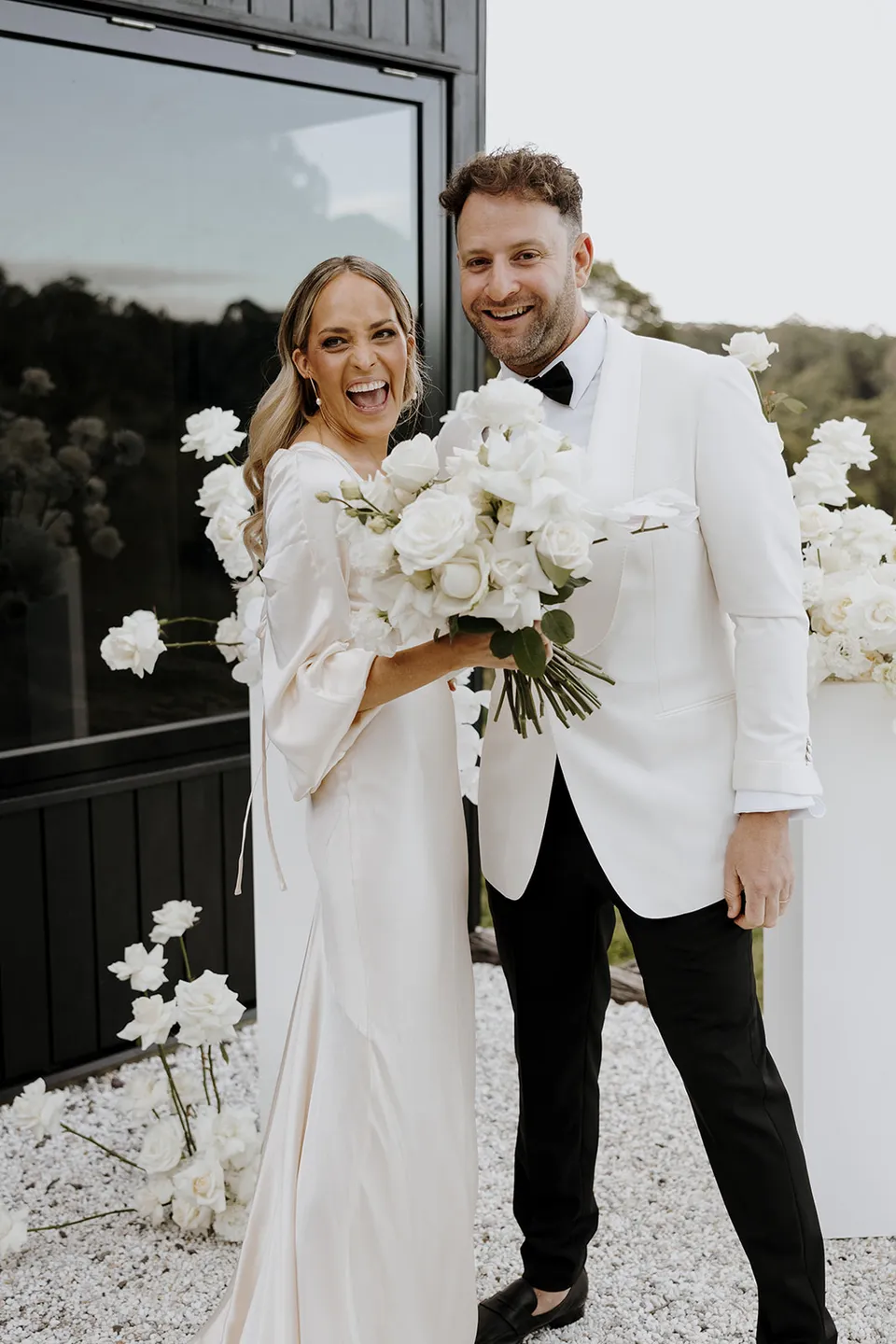 Gabrielle and Greg smiling together with her white bouquet beside the cabin after their ceremony at Roundhill Retreat in Obi Obi, Sunshine Coast Hinterland, for their Elopement Collective elopement.
