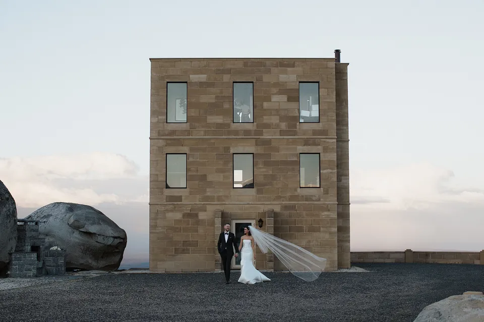 Meg and Ryan standing in front of the sandstone tower beneath a soft evening sky, with her veil stretching in the breeze. At The Keep on Tasmania’s east coast, for an Elopement Collective elopement. Photograph by Jenna Agius.