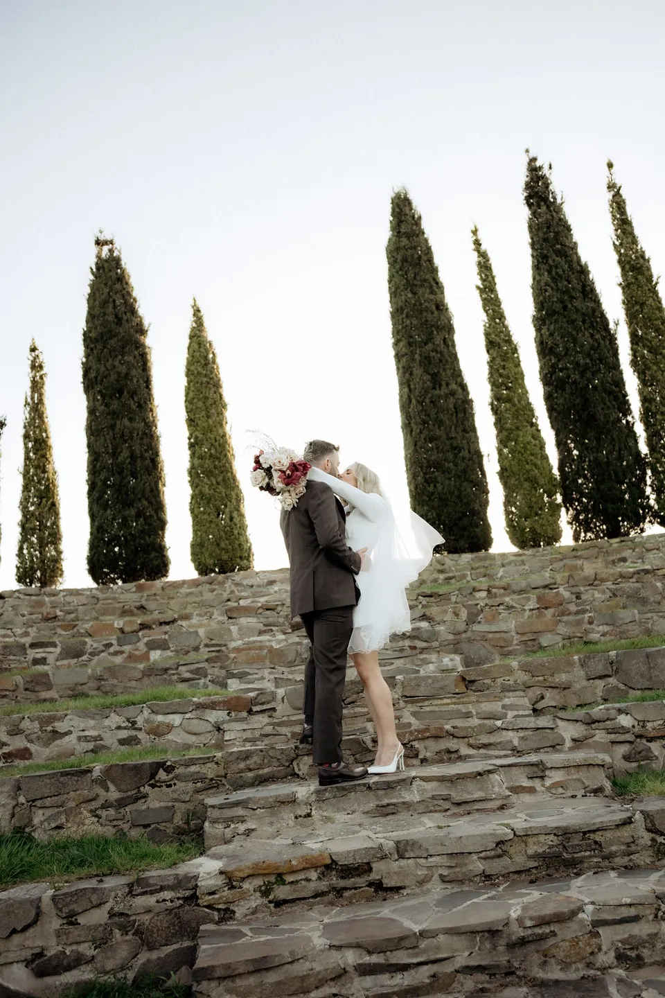 Kaitlyn and Russell kissing on the stone steps, with her veil lifted and bouquet by The Romantics raised beside them. At Piermont Retreat on Tasmania’s east coast, for an Elopement Collective elopement. Photograph by Omar of By The Wilde.