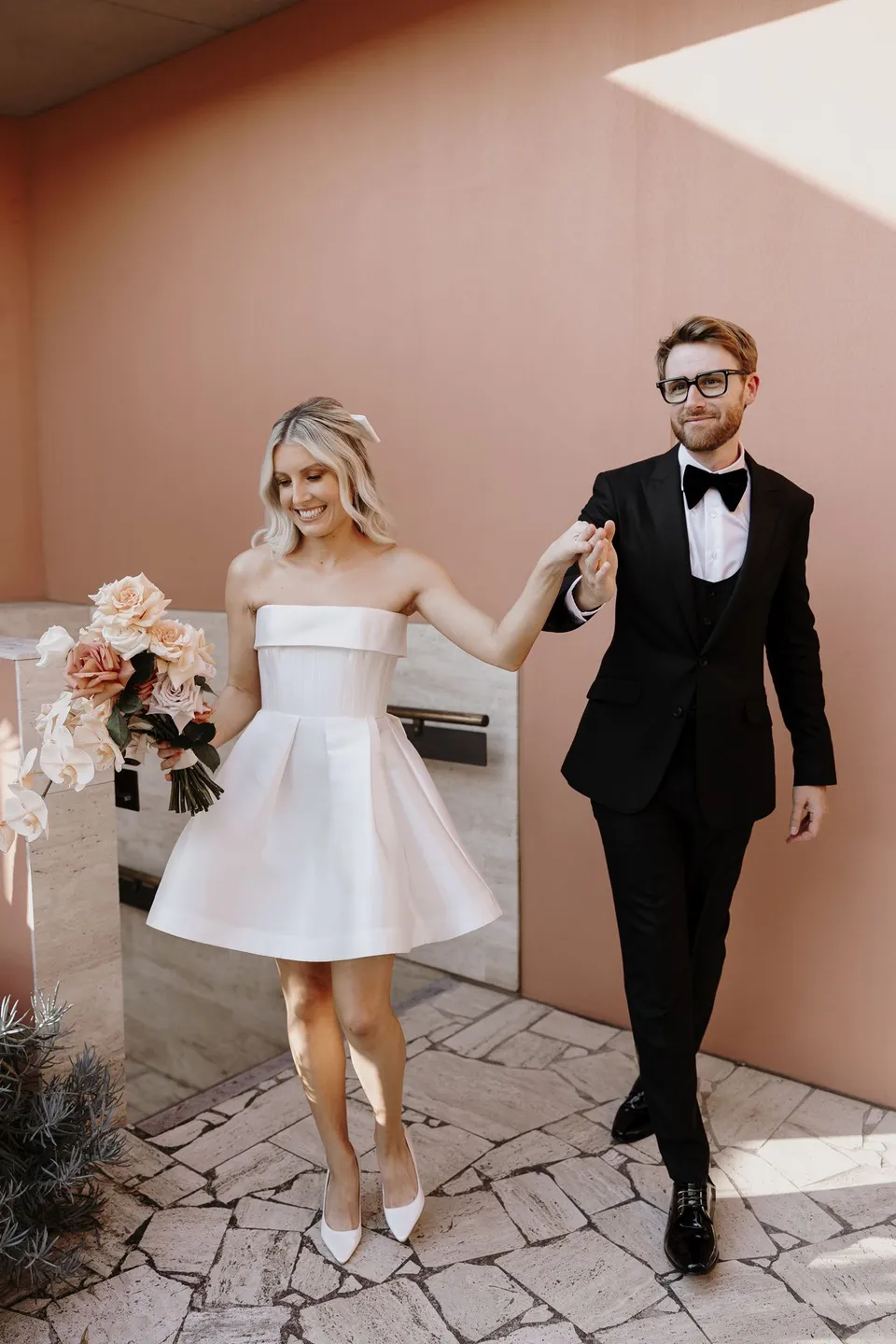 Danielle and Adam walking hand in hand across the stone terrace beside a pink wall, during Danielle and Adam’s Elopement Collective elopement at The Calile Hotel in Fortitude Valley, Brisbane.