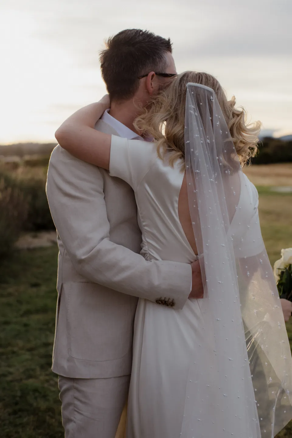The couple embracing beside the stone house with Jemmah’s veil across her back, at Thalia Haven on the East Coast of Tasmania, Australia, for an Elopement Collective elopement. Photograph by Joey Willis.