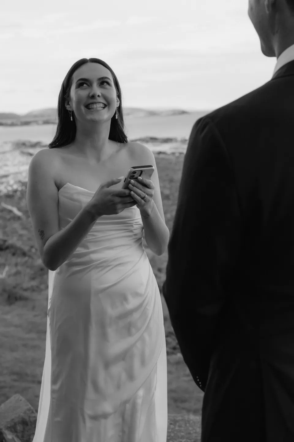 Black and white portrait of Dani smiling at Anthony while holding her phone to read her personal vows, at Thalia Haven on Tasmania’s east coast, for an Elopement Collective elopement. Photograph by Bulb Creative.