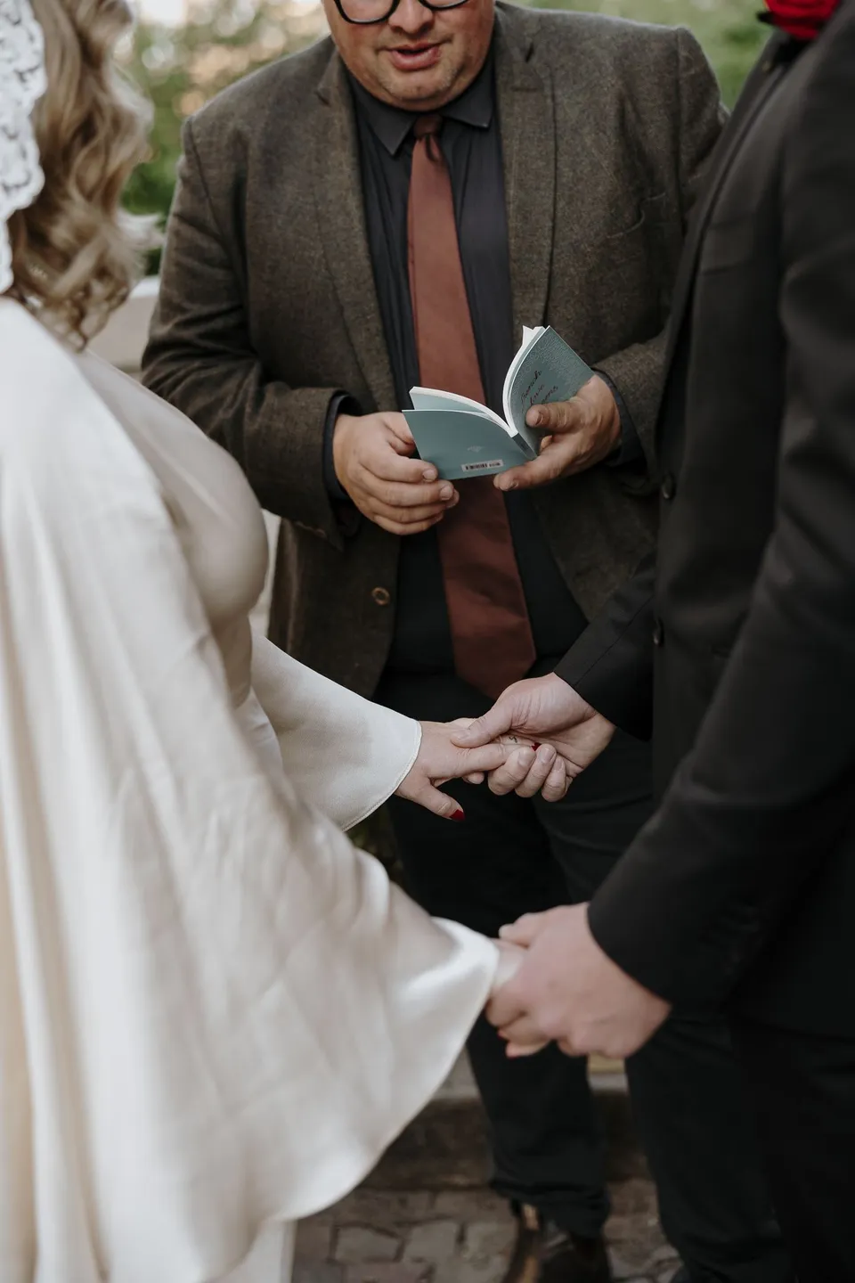 Close view of Kara and Samuel holding hands during their ceremony, with celebrant Josh Withers holding an open book behind them, at Avenue de Camoëns in Paris, France, during an Elopement Collective elopement. Photograph by George Bowden.