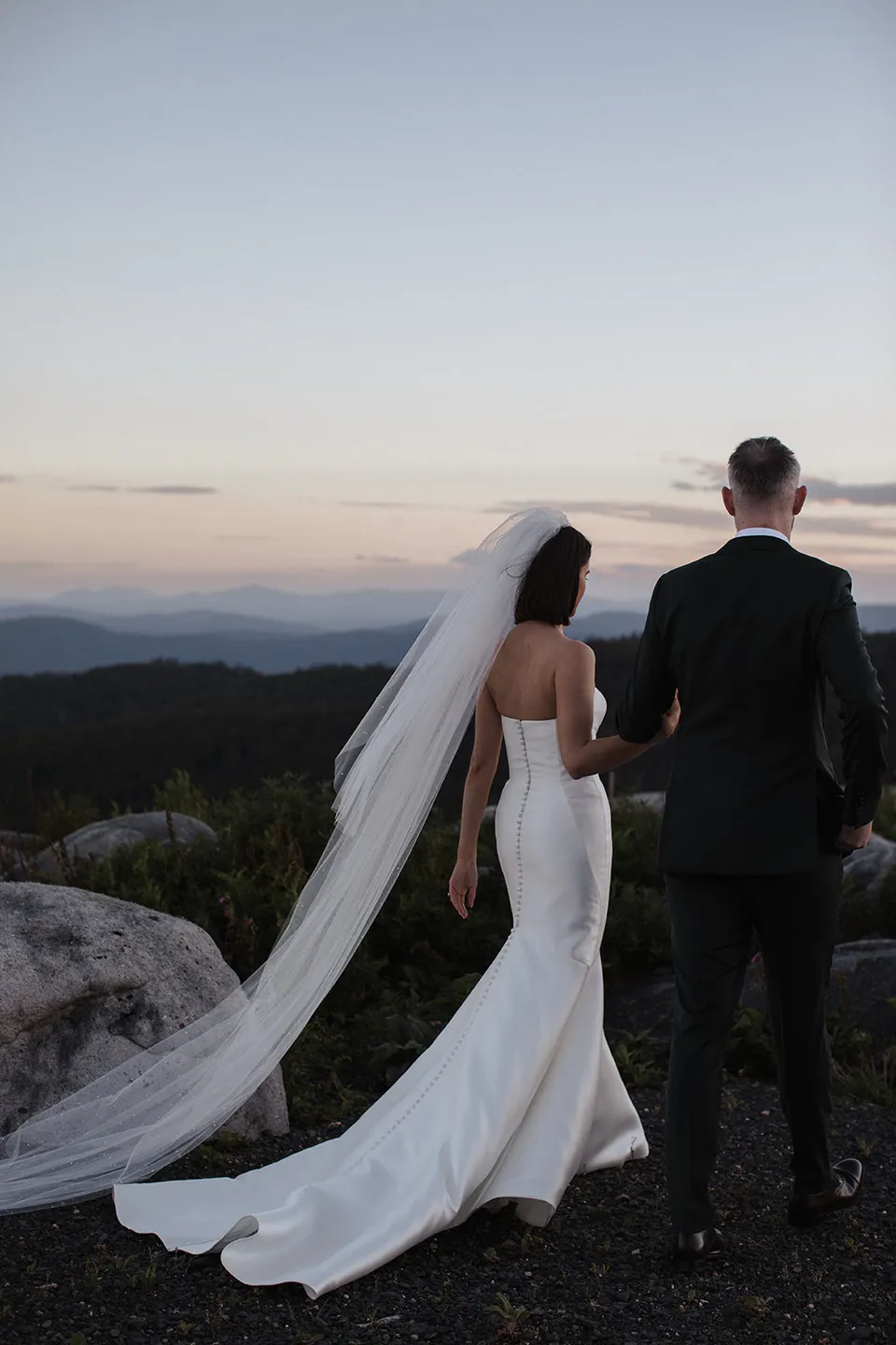 Meg and Ryan looking out across forested valleys, with her veil and dress trailing across the rock behind them. At The Keep on Tasmania’s east coast, for an Elopement Collective elopement. Photograph by Jenna Agius.