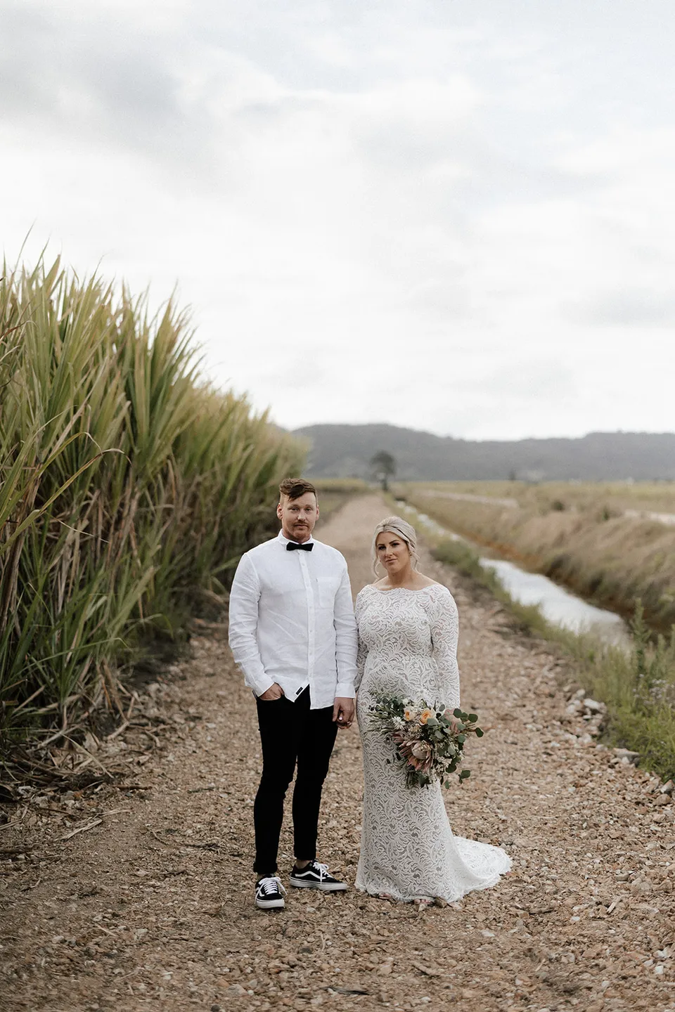 Matt and Laura standing beside a cane field and narrow waterway, with Laura holding her Fifth Avenue Florist bouquet, in the Tweed Valley, NSW, for an Elopement Collective elopement. Photo by Bulb Creative.