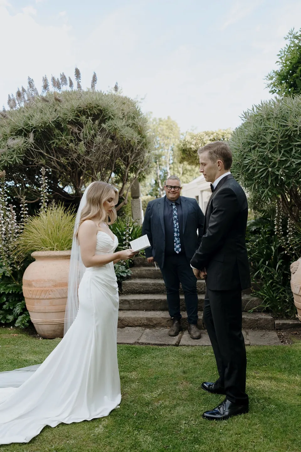 Tanesha reading her vows to Andrew while celebrant Josh Withers smiles behind them beside the garden steps, at Islington Hotel in South Hobart, Tasmania, Australia, for an Elopement Collective summer elopement. Photograph by George Bowden.