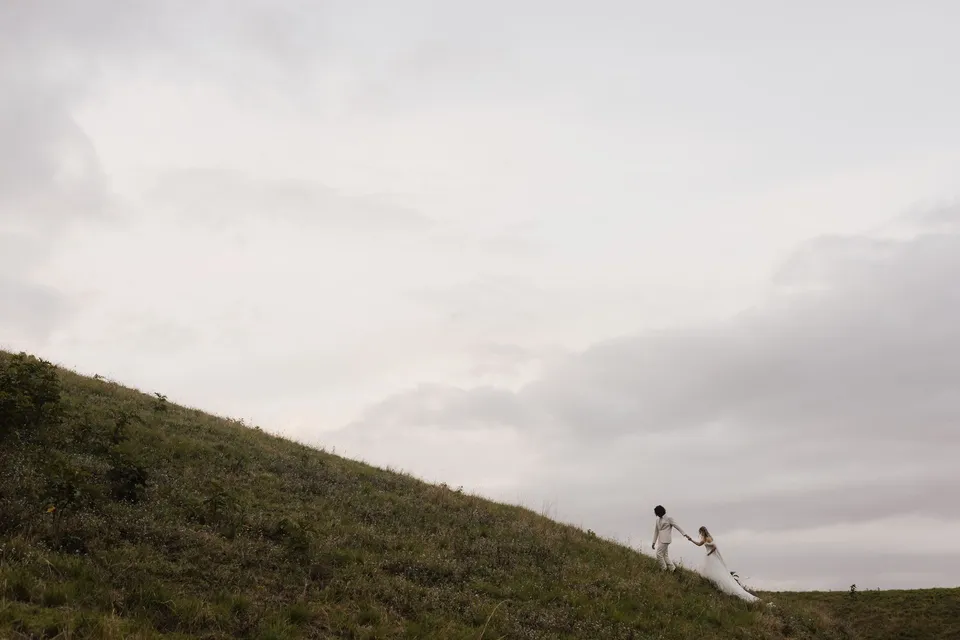 Bridget and James running hand in hand along the hillside beneath a wide sky. At Cudgera Creek in northern New South Wales, for an Elopement Collective elopement. Photograph by Bulb Creative.
