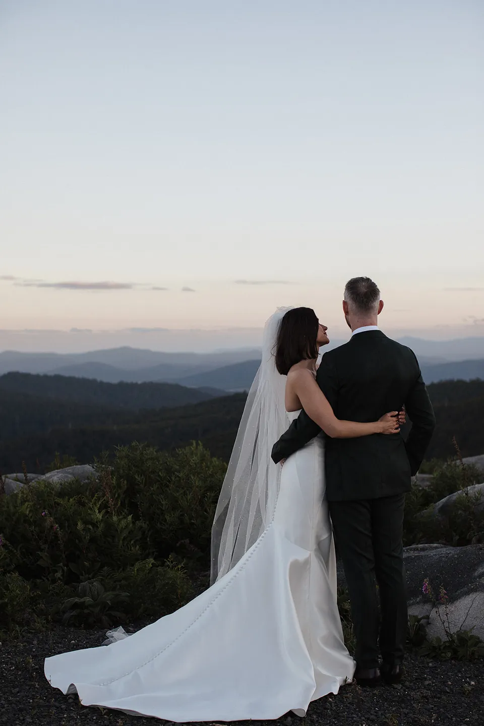 Meg and Ryan with their arms around each other, looking across layers of hills beneath a pale evening sky. At The Keep on Tasmania’s east coast, for an Elopement Collective elopement. Photograph by Jenna Agius.