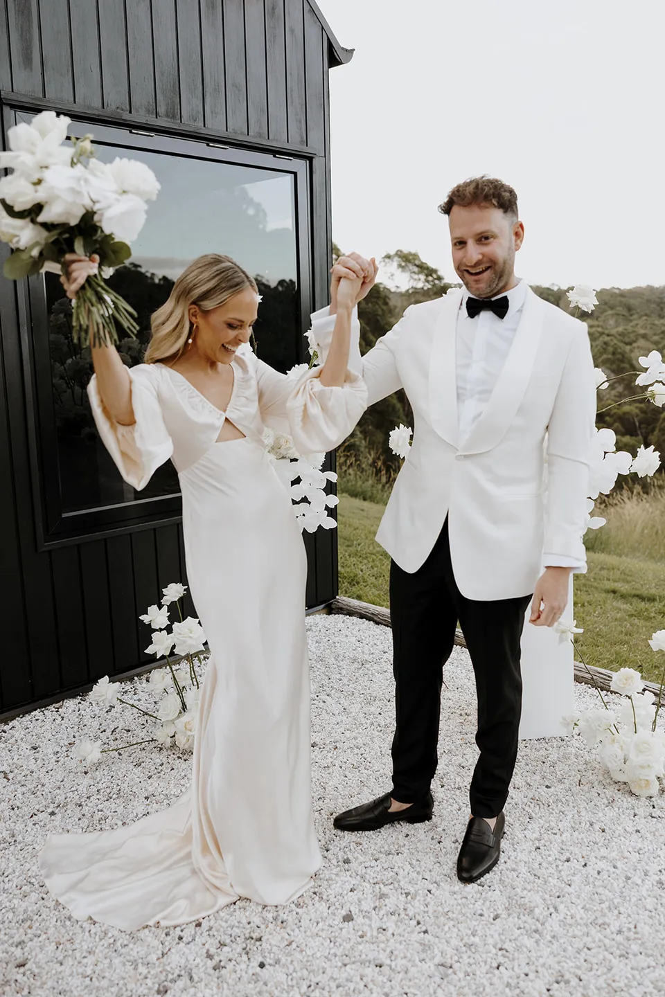 Gabrielle lifting her bouquet as Greg raises their joined hands in celebration outside the cabin at Roundhill Retreat in Obi Obi, Sunshine Coast Hinterland, for their Elopement Collective elopement.
