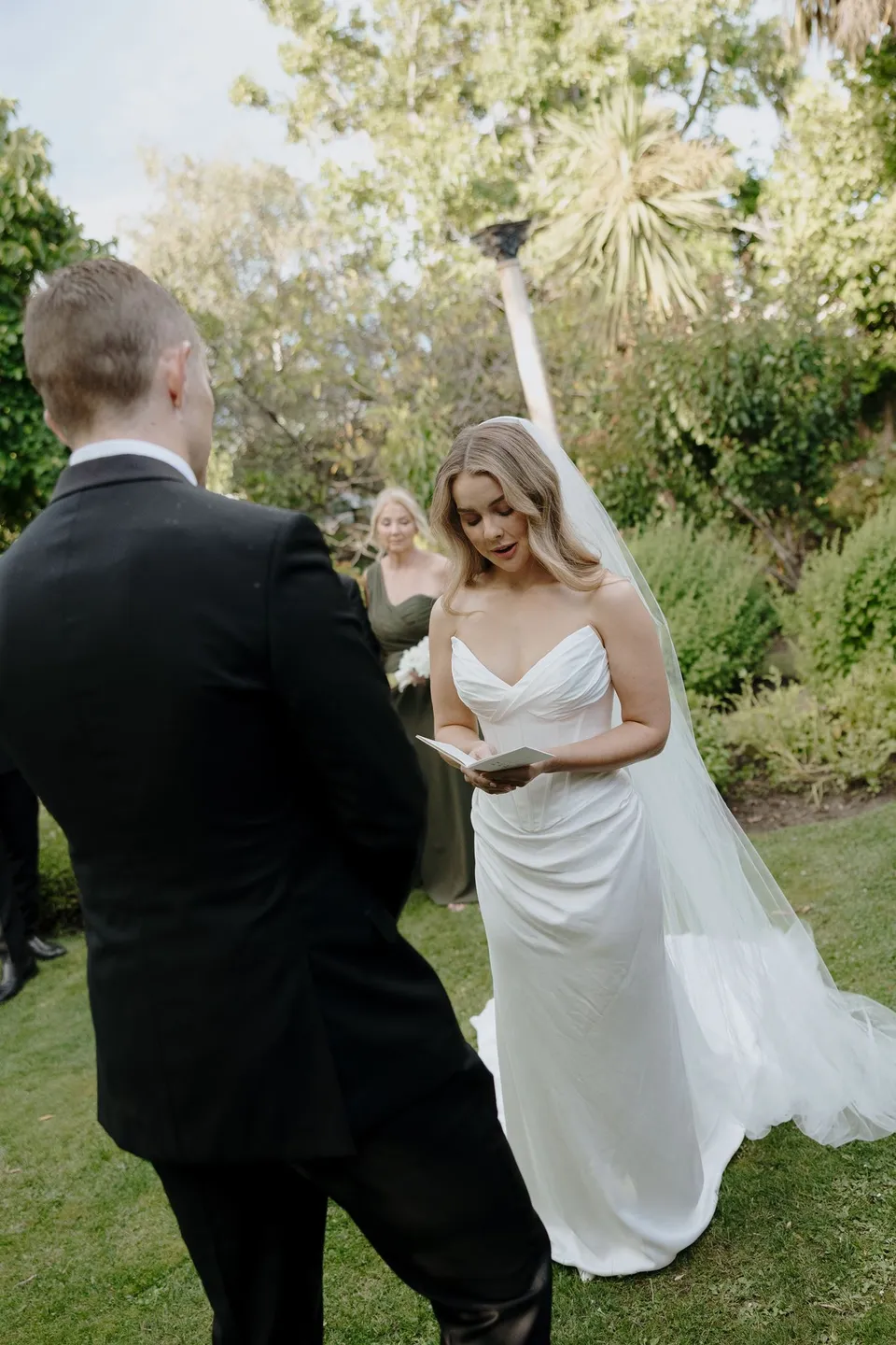 Tanesha lifting a hand to her eye while standing opposite Andrew during their garden ceremony, at Islington Hotel in South Hobart, Tasmania, Australia, for an Elopement Collective summer elopement. Photograph by George Bowden.