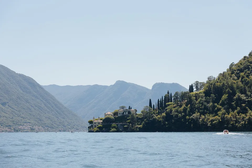 Villa del Balbianello seen from the water on its wooded peninsula, with the mountains rising behind it, at Lake Como in Lombardy, Italy, for an Elopement Collective elopement. Photograph by George Bowden.