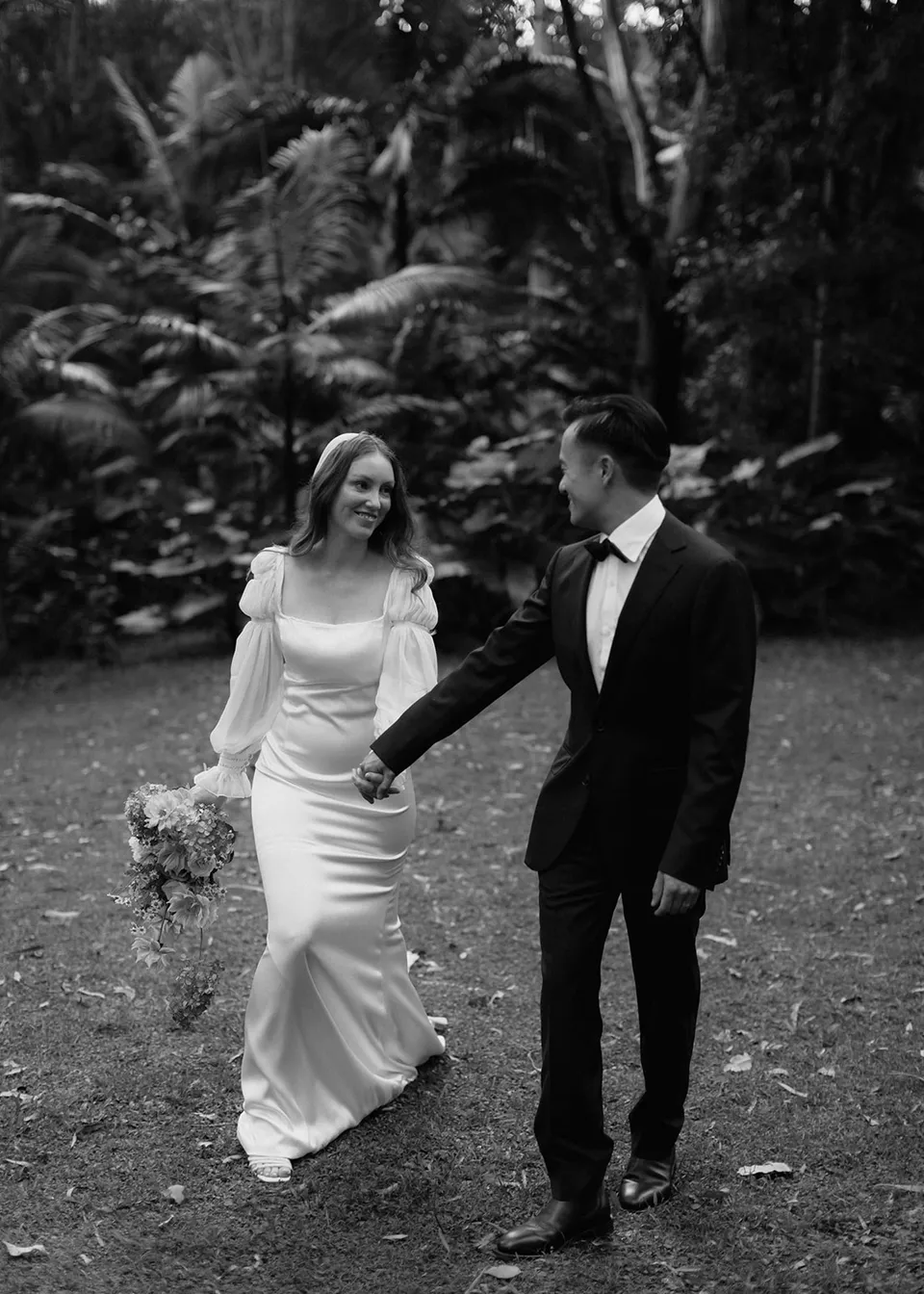 Black-and-white photograph of the couple walking together with their hands joined. At Fig Tree Villa in Ewingsdale, Byron Bay, for an Elopement Collective elopement. Photograph by Bulb Creative.
