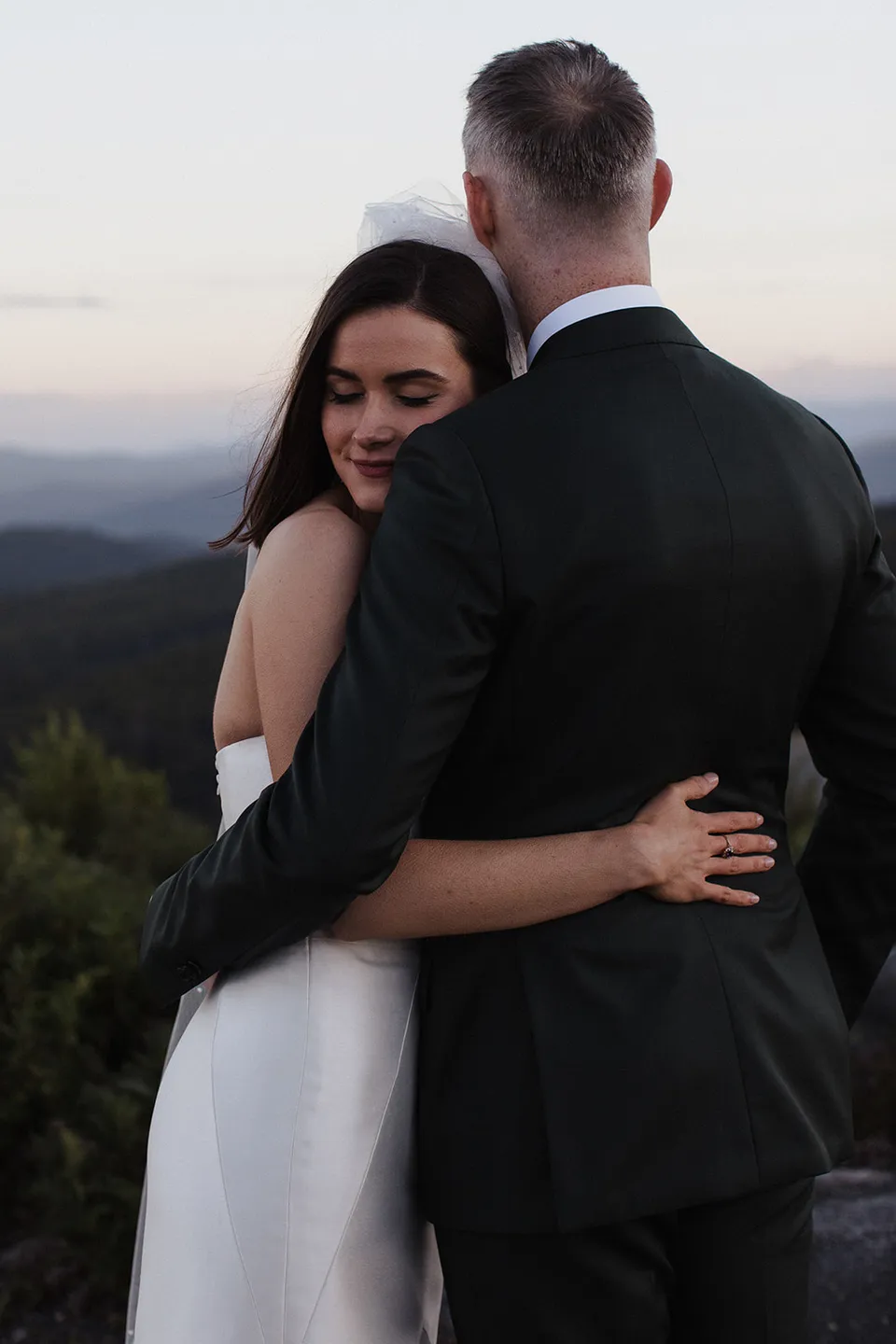 Meg resting against Ryan with her eyes closed as they embrace above the forest. At The Keep on Tasmania’s east coast, for an Elopement Collective elopement. Photograph by Jenna Agius.