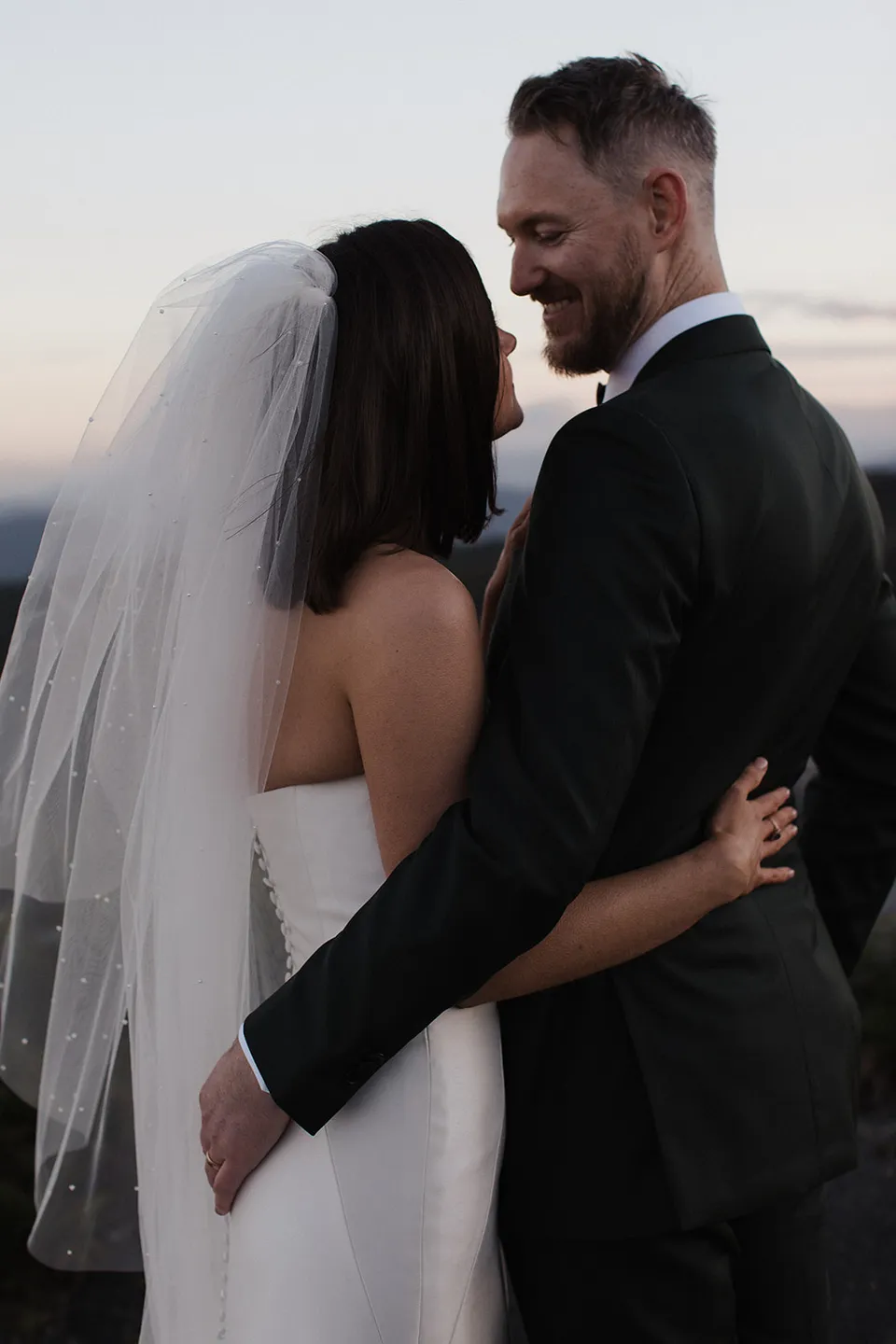 Ryan smiling at Meg as they stand with their arms around each other on the hilltop. At The Keep on Tasmania’s east coast, for an Elopement Collective elopement. Photograph by Jenna Agius.