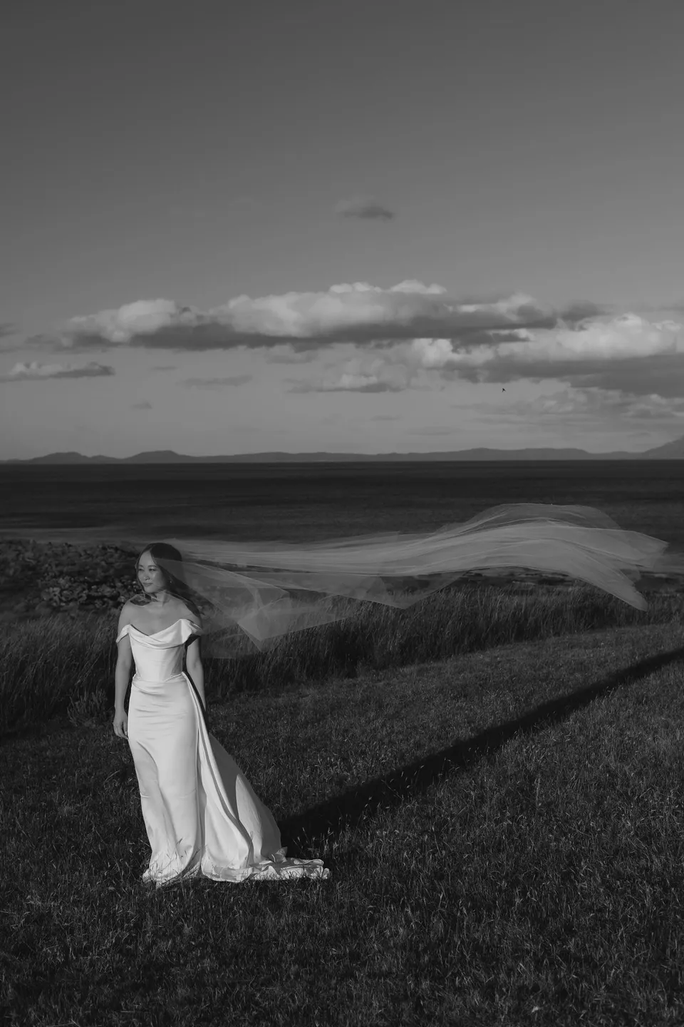 Black and white portrait of Clara in her white dress among coastal grasses, with the bay behind her, at Thalia Haven on the East Coast of Tasmania, for an Elopement Collective elopement. Photograph by Bulb Creative.
