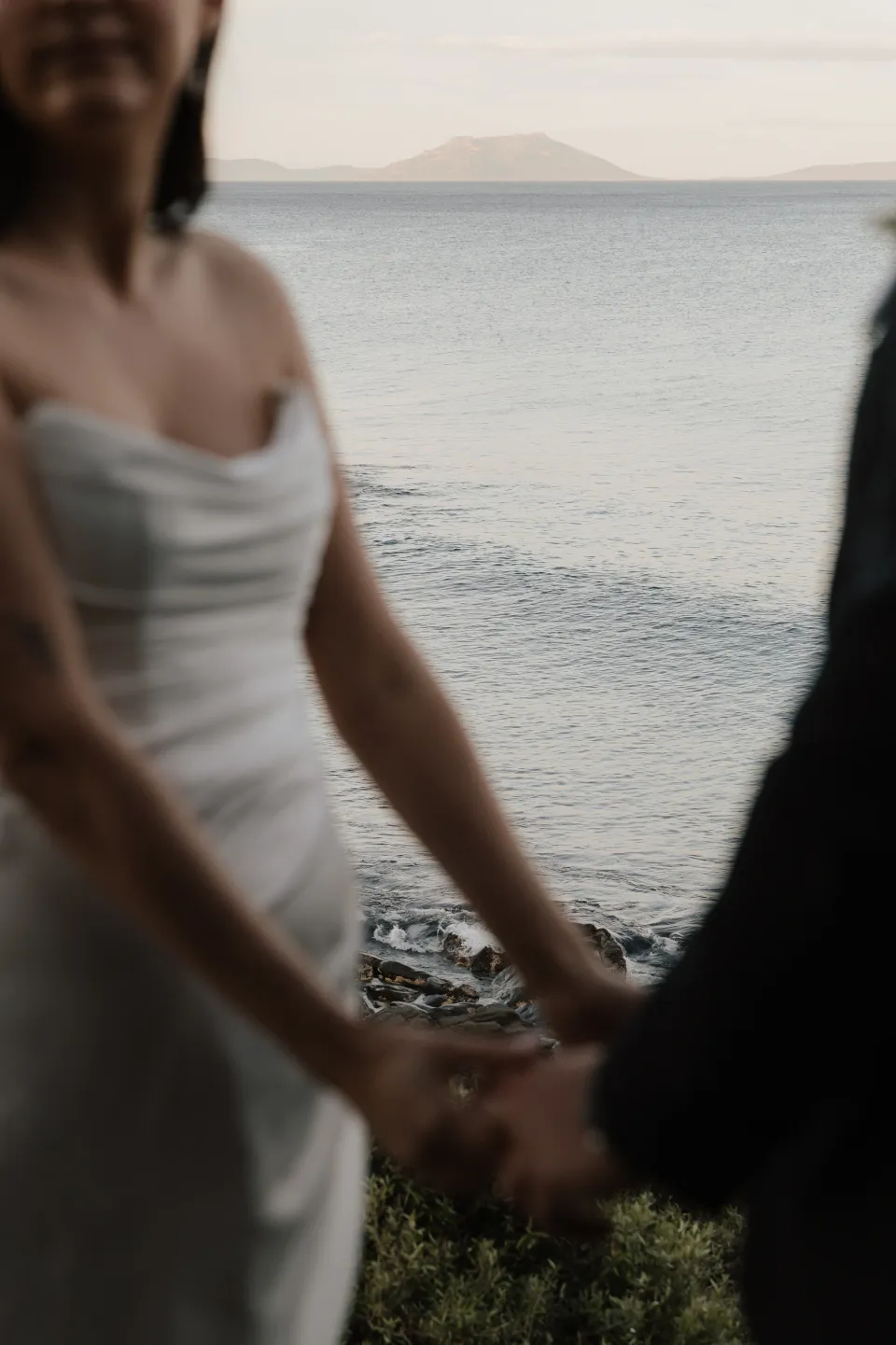 Close view of Dani and Anthony holding hands on the rocky shoreline, with blue-grey water behind them, at Thalia Haven on Tasmania’s east coast, for an Elopement Collective elopement. Photograph by Bulb Creative.