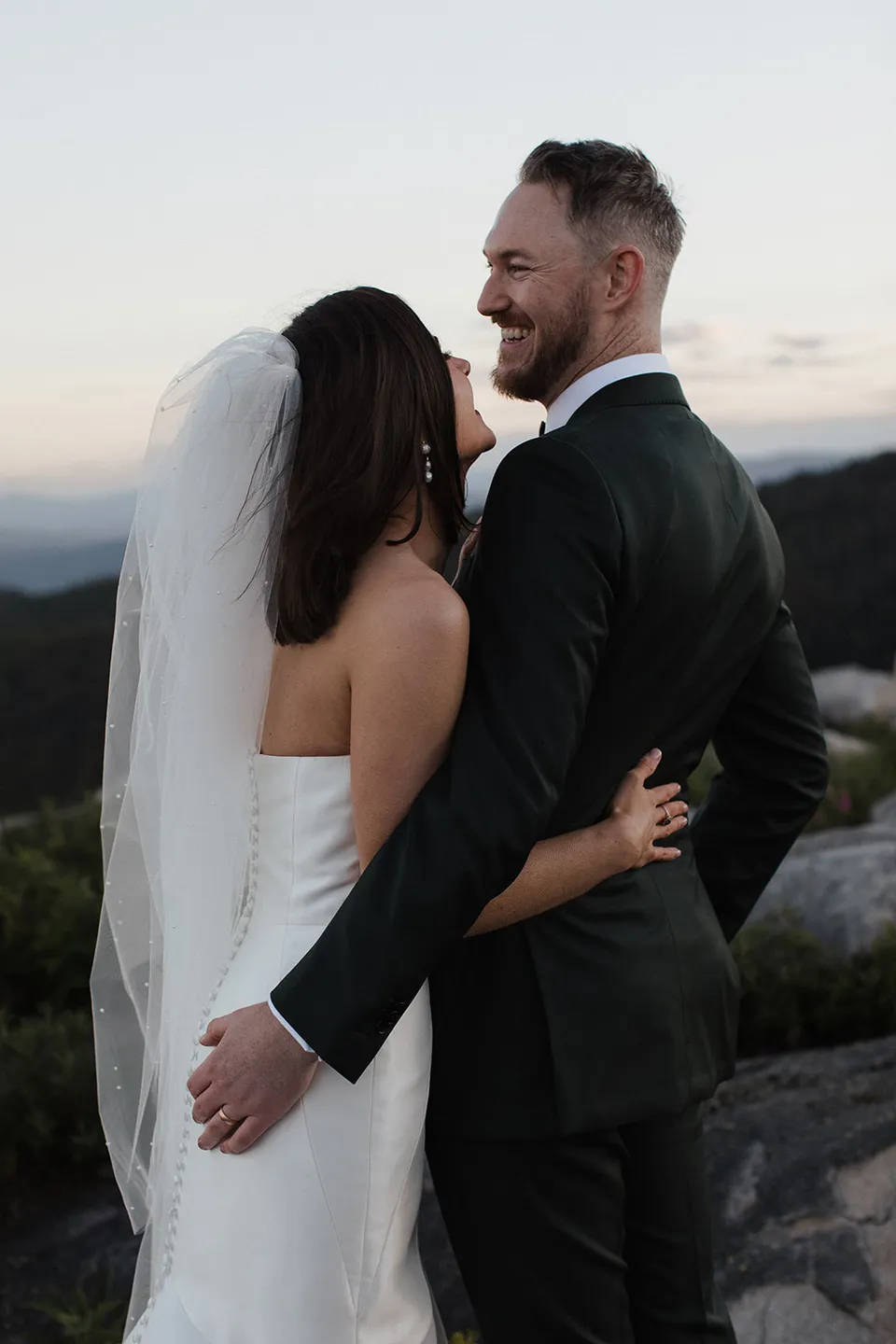 Meg and Ryan sharing a laugh in an embrace, with her veil falling down her back. At The Keep on Tasmania’s east coast, for an Elopement Collective elopement. Photograph by Jenna Agius.