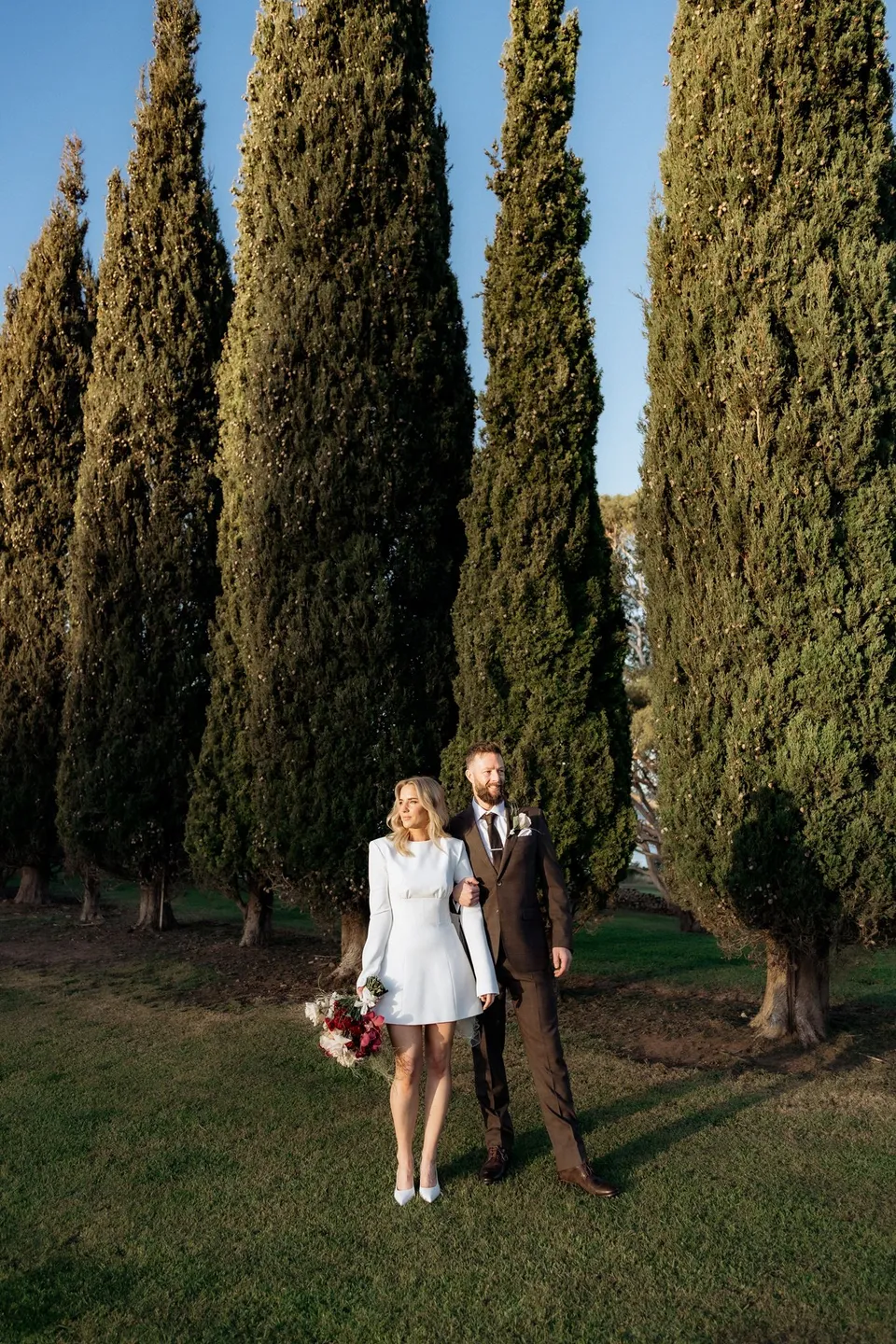 Kaitlyn and Russell standing together beneath tall cypress trees, with her bouquet by The Romantics at her side. At Piermont Retreat on Tasmania’s east coast, for an Elopement Collective elopement. Photograph by Omar of By The Wilde.