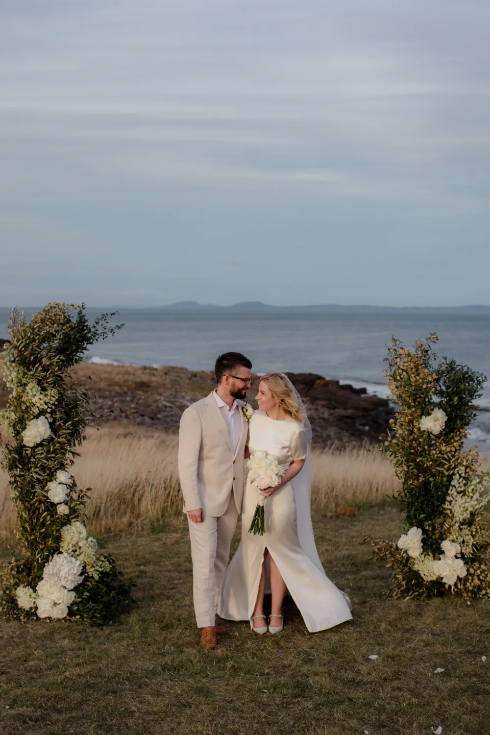 The couple leaning close beside a ceremony flower arrangement by The Romantics, at Thalia Haven on the East Coast of Tasmania, Australia, for an Elopement Collective elopement. Photograph by Joey Willis.