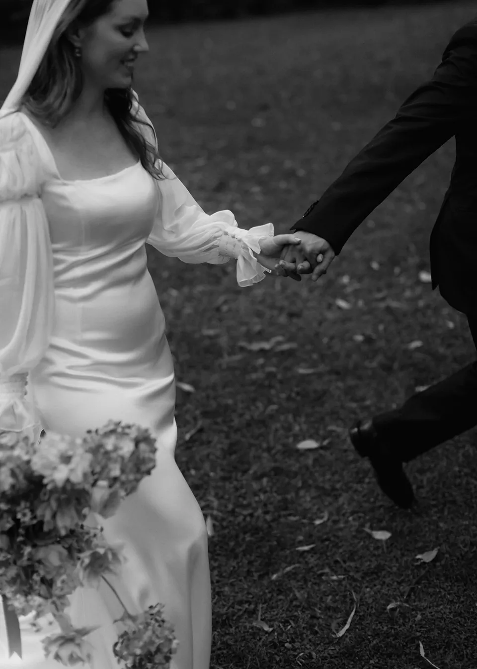 Black-and-white detail of the couple's joined hands beside Elyse's bouquet, with flowers by The Perfect Party Co. At Fig Tree Villa in Ewingsdale, Byron Bay, for an Elopement Collective elopement. Photograph by Bulb Creative.