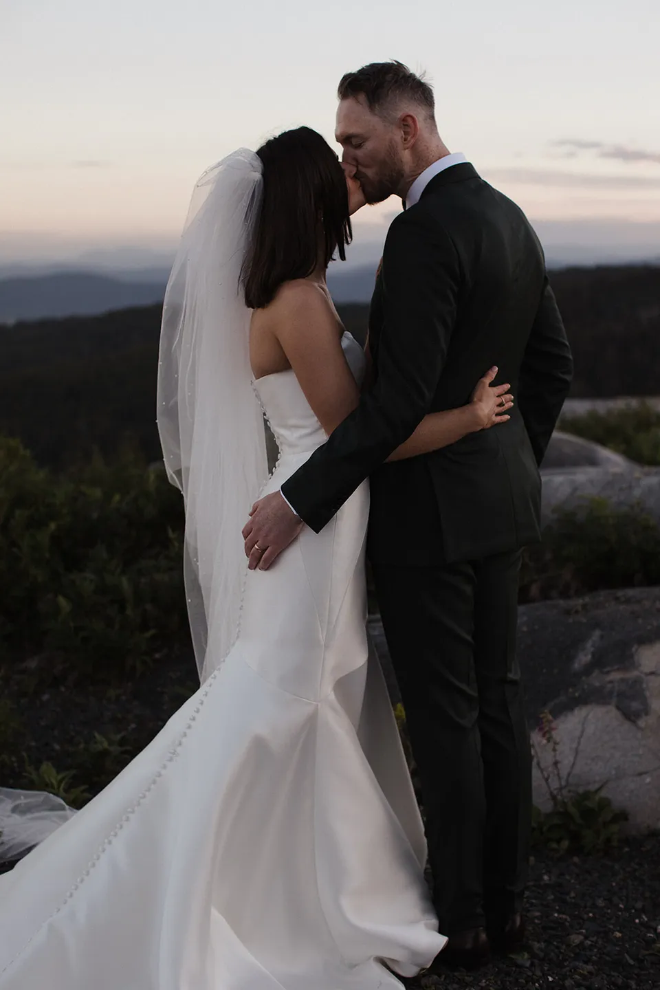 Meg and Ryan standing close together above the forest as the evening sky softens behind them. At The Keep on Tasmania’s east coast, for an Elopement Collective elopement. Photograph by Jenna Agius.