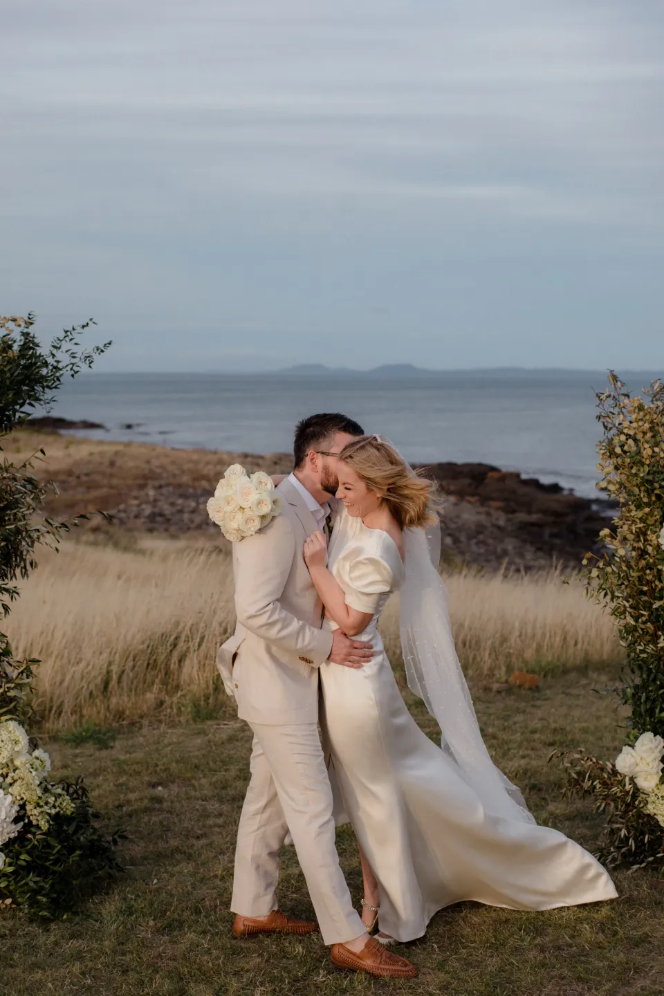 The couple embracing between the ceremony flowers with the coast behind them, at Thalia Haven on the East Coast of Tasmania, Australia, for an Elopement Collective elopement. Photograph by Joey Willis.