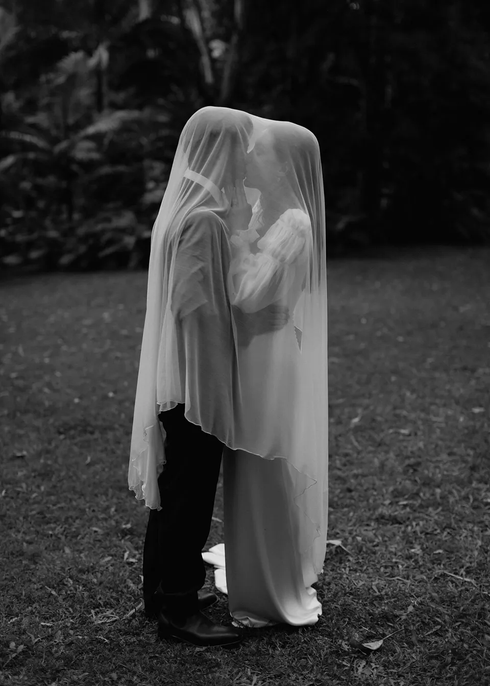 Black-and-white full-length portrait of the couple standing together beneath Elyse's veil. At Fig Tree Villa in Ewingsdale, Byron Bay, for an Elopement Collective elopement. Photograph by Bulb Creative.