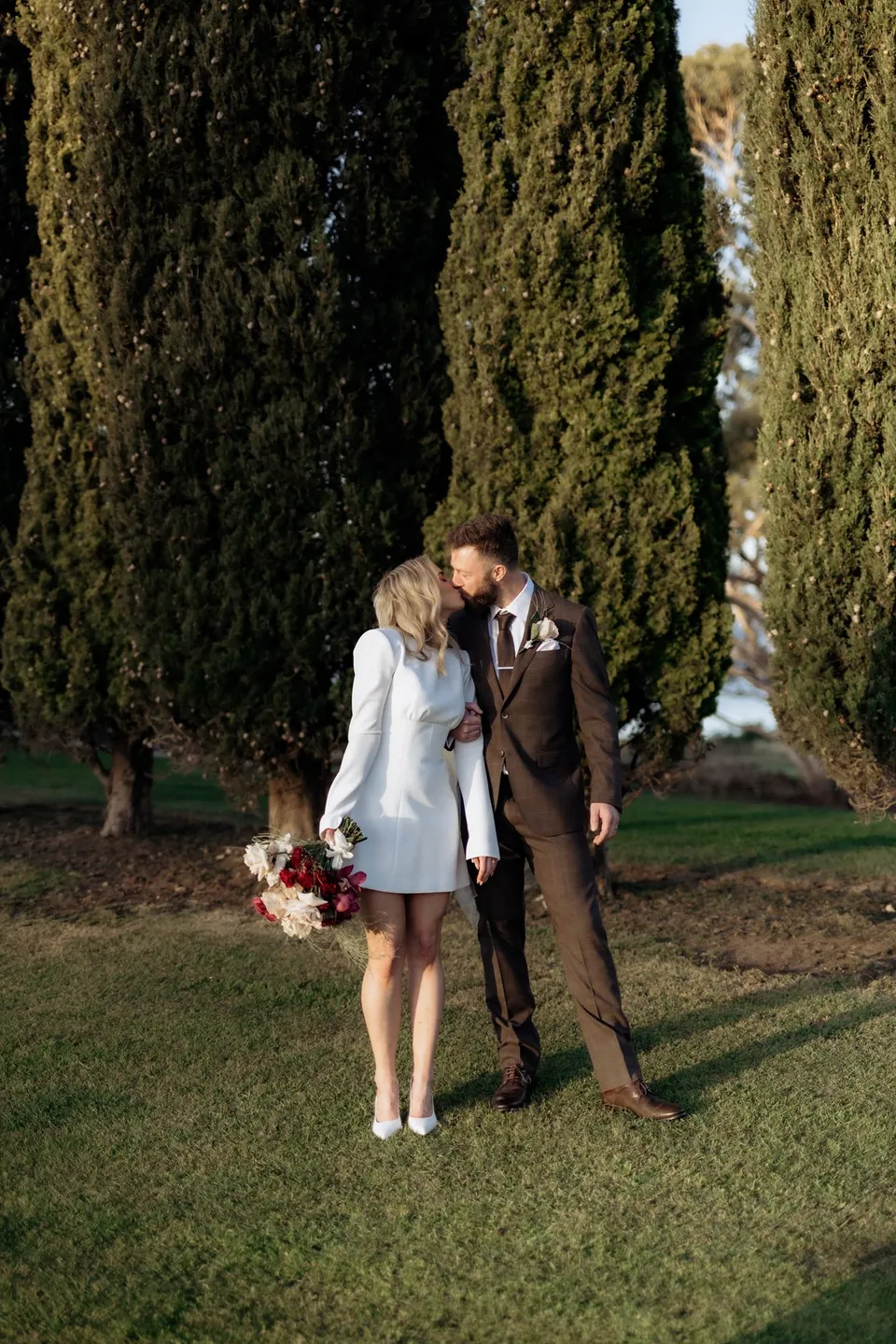 Russell kissing Kaitlyn’s forehead beneath the cypress trees as she holds her bouquet by The Romantics. At Piermont Retreat on Tasmania’s east coast, for an Elopement Collective elopement. Photograph by Omar of By The Wilde.