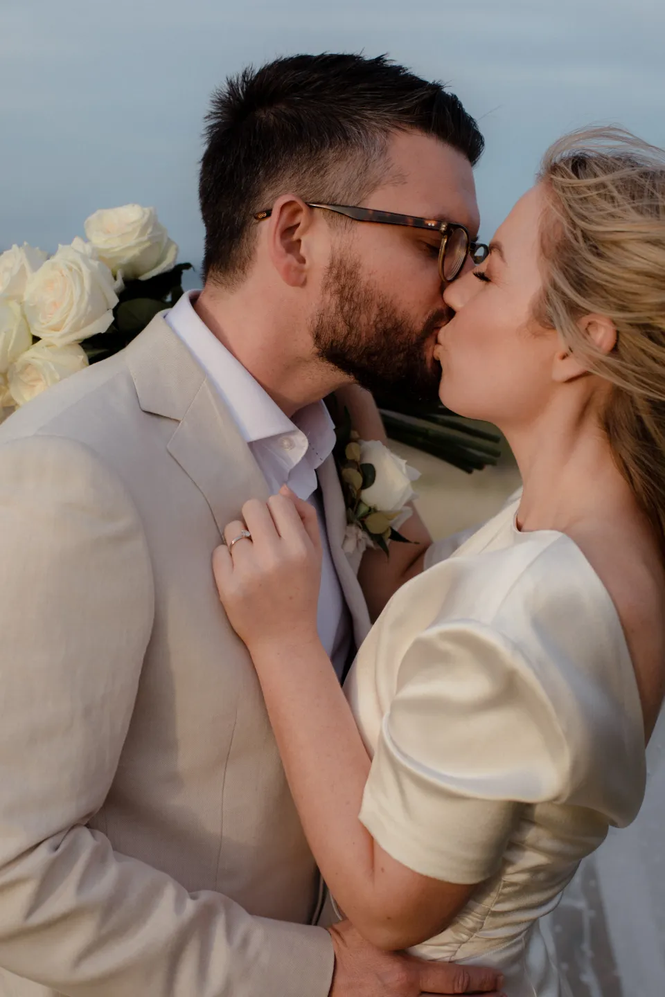 A close portrait of Jemmah kissing Jack with her white bouquet by The Romantics, at Thalia Haven on the East Coast of Tasmania, Australia, for an Elopement Collective elopement. Photograph by Joey Willis.