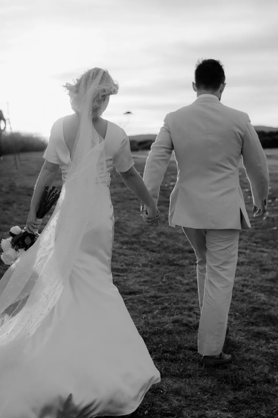 Black and white rear view of the newlyweds walking hand in hand, at Thalia Haven on the East Coast of Tasmania, Australia, for an Elopement Collective elopement. Photograph by Joey Willis.