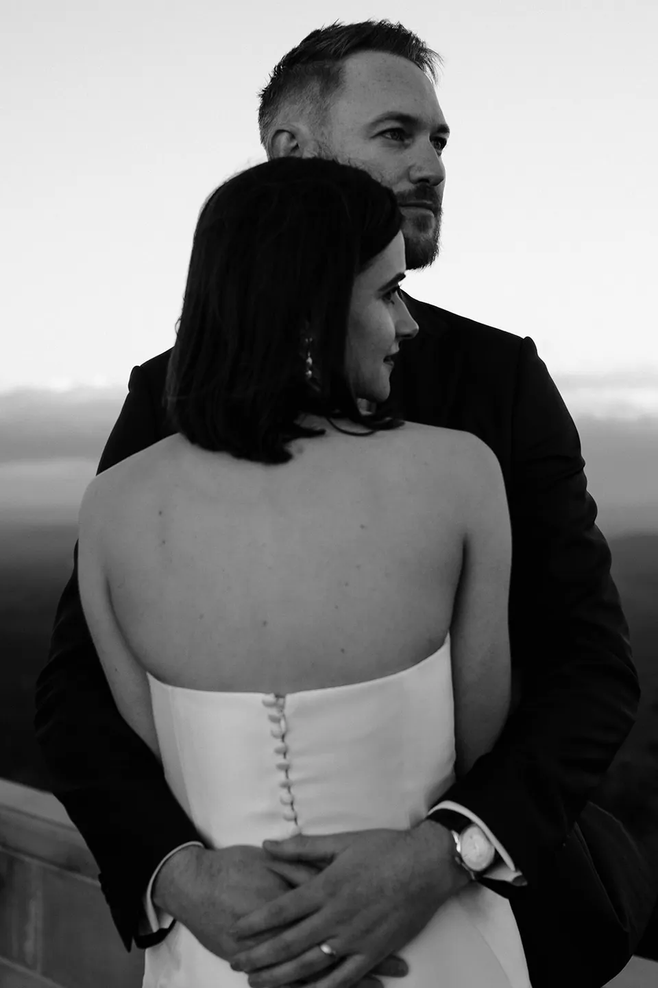Black and white portrait of Ryan holding Meg, showing the line of buttons down the back of her dress. At The Keep on Tasmania’s east coast, for an Elopement Collective elopement. Photograph by Jenna Agius.