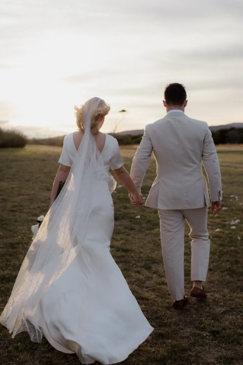 Jemmah and Jack walking away with her veil and dress trailing behind, at Thalia Haven on the East Coast of Tasmania, Australia, for an Elopement Collective elopement. Photograph by Joey Willis.