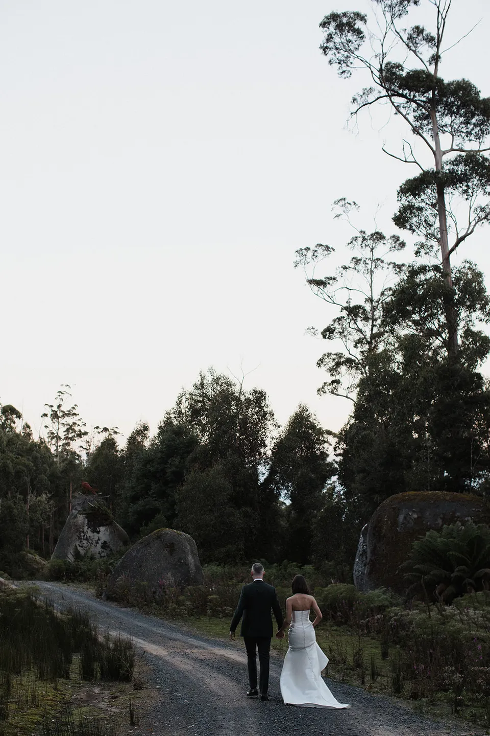 Meg and Ryan walking away along a gravel drive bordered by tall trees and ferns. At The Keep on Tasmania’s east coast, for an Elopement Collective elopement. Photograph by Jenna Agius.