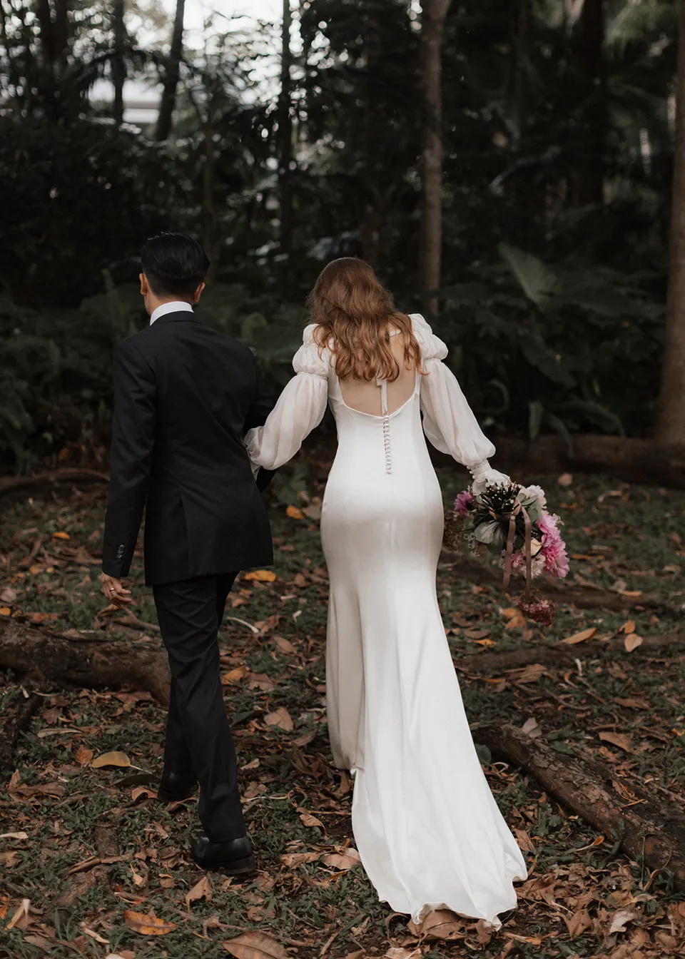 The couple strolling towards the trees with Elyse's long train behind her. At Fig Tree Villa in Ewingsdale, Byron Bay, for an Elopement Collective elopement. Photograph by Bulb Creative.