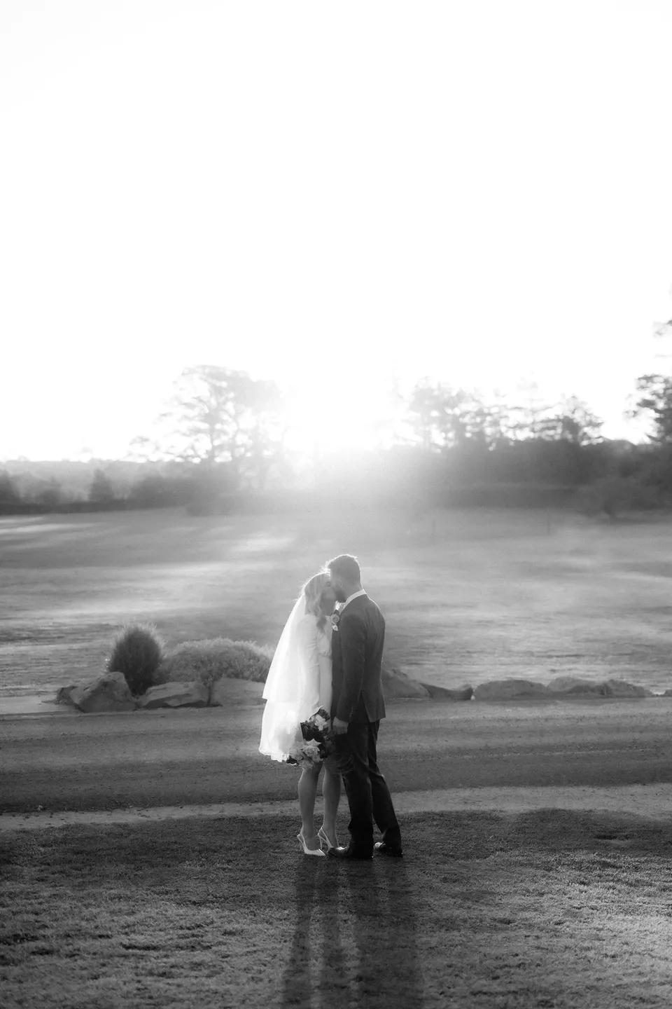 Black and white portrait of Kaitlyn and Russell embracing, with low sunlight casting long shadows across the lawn. At Piermont Retreat on Tasmania’s east coast, for an Elopement Collective elopement. Photograph by Omar of By The Wilde.