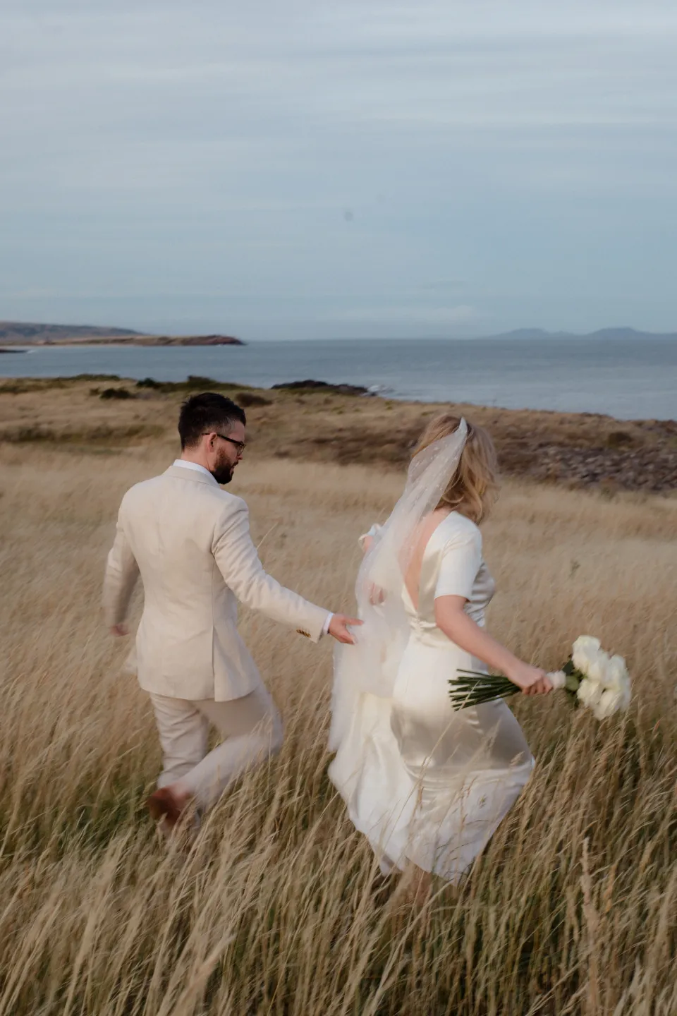 The couple walking through tall coastal grasses with Jemmah’s bouquet by The Romantics, at Thalia Haven on the East Coast of Tasmania, Australia, for an Elopement Collective elopement. Photograph by Joey Willis.