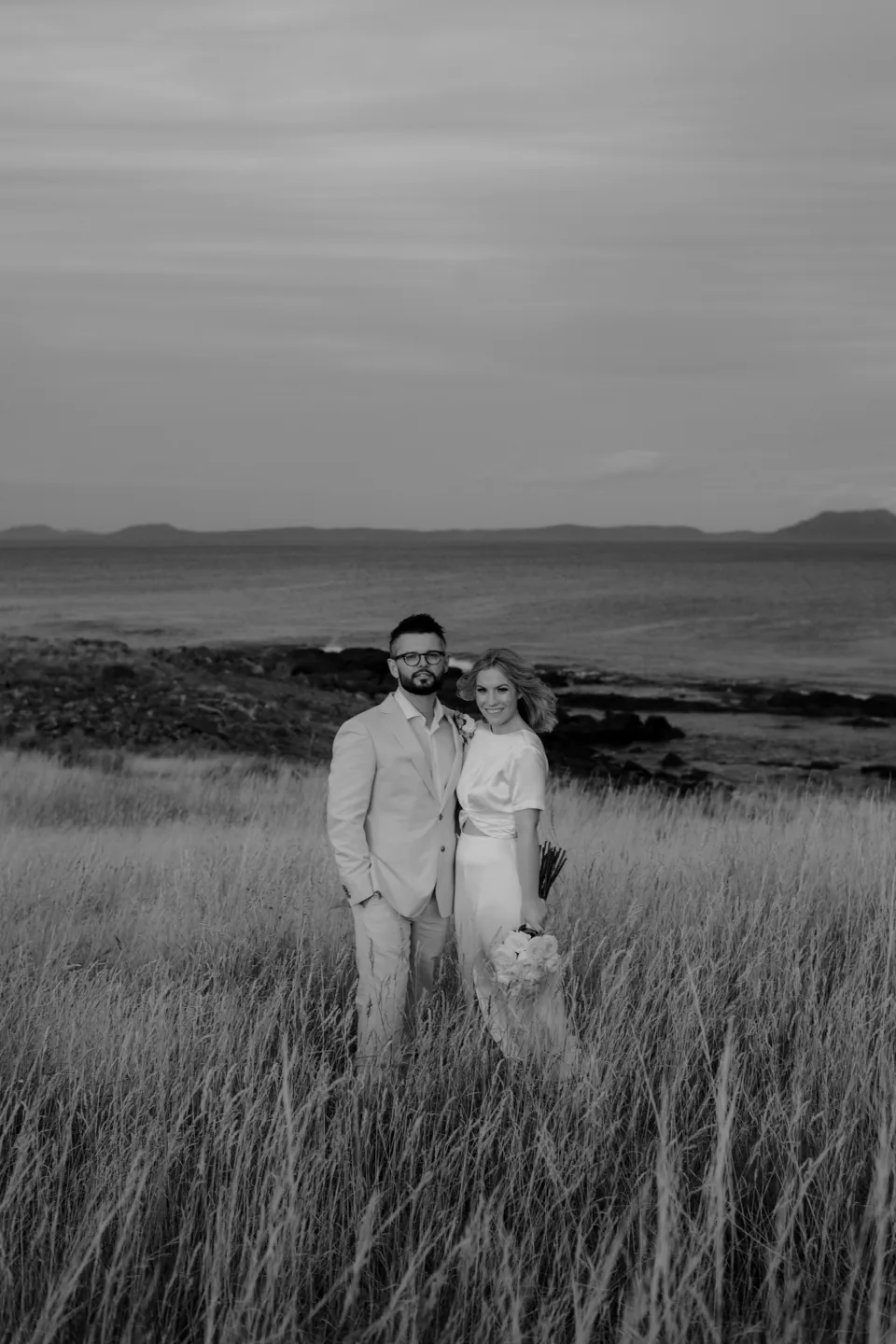 Black and white portrait of the couple standing close in tall grass, at Thalia Haven on the East Coast of Tasmania, Australia, for an Elopement Collective elopement. Photograph by Joey Willis.