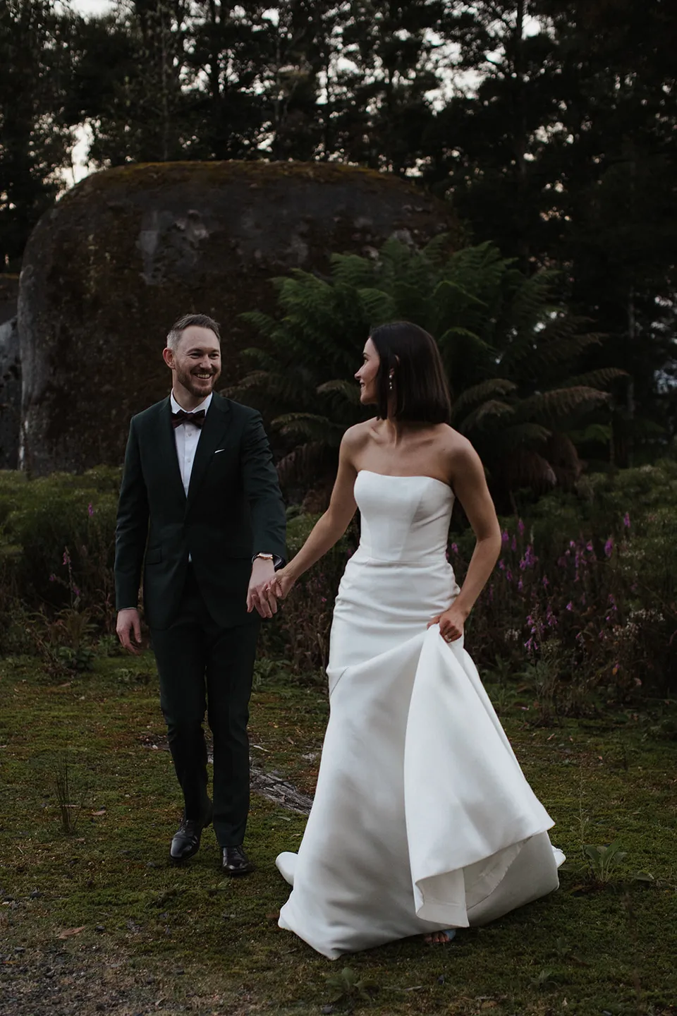Meg looking back at Ryan as they laugh and walk hand in hand along a grassy woodland path. At The Keep on Tasmania’s east coast, for an Elopement Collective elopement. Photograph by Jenna Agius.