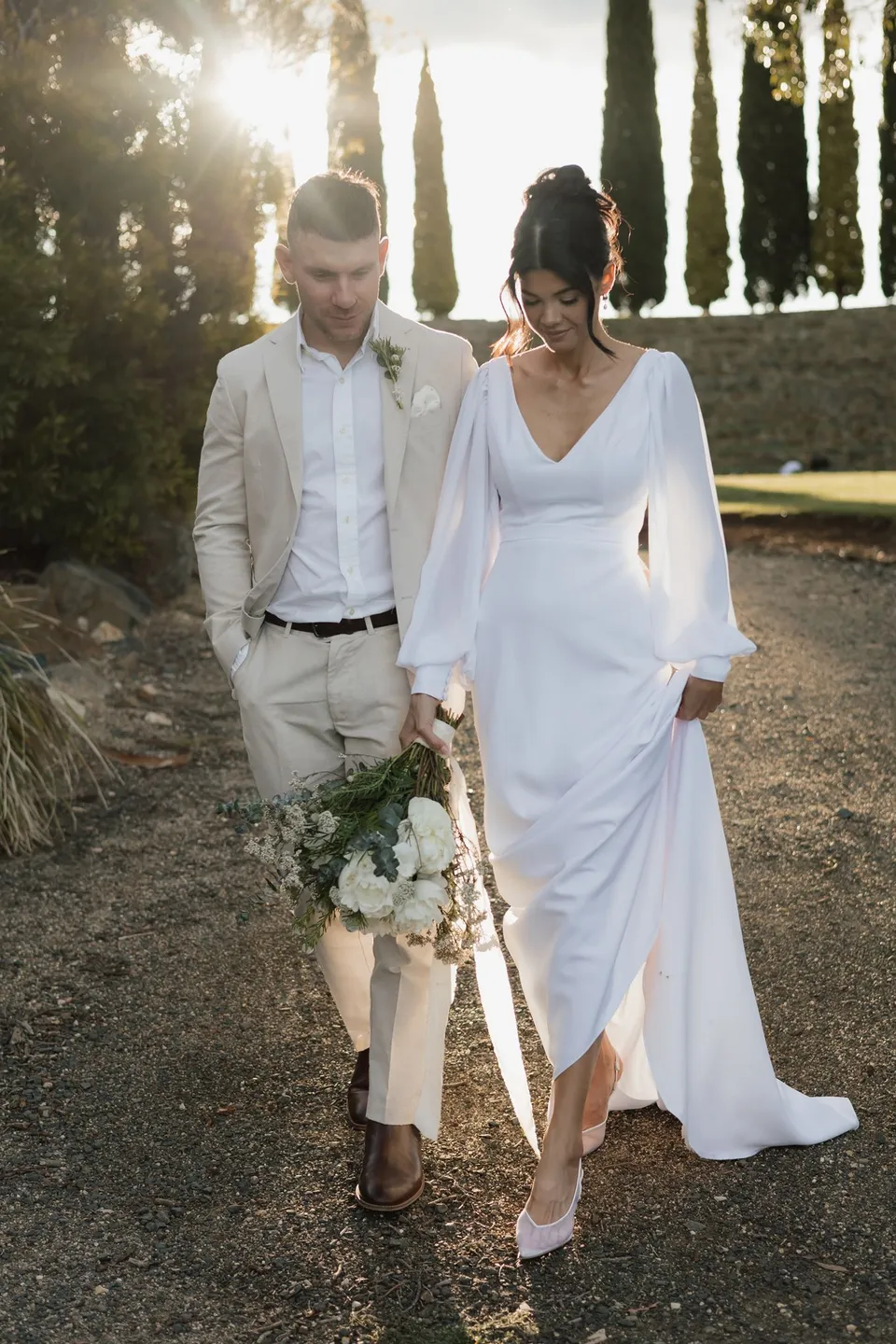 Renee and Mitch walking together along a gravel path in low sunlight, carrying her white bouquet by The Romantics. At Piermont Retreat on Tasmania’s east coast, for an Elopement Collective elopement. Photograph by Bulb Creative.