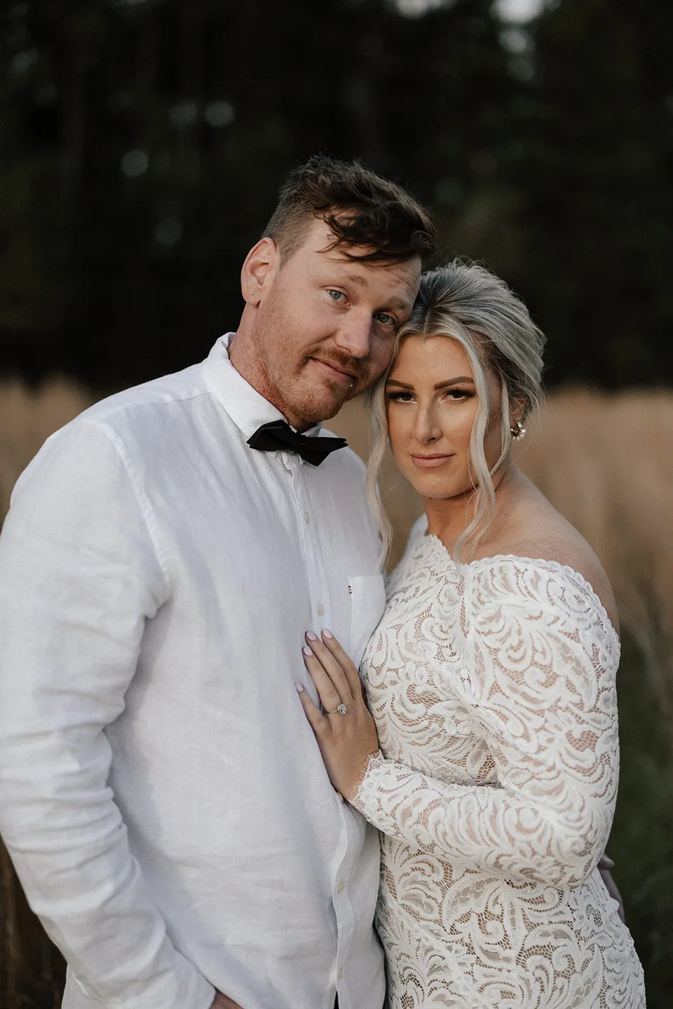 Matt resting his head against Laura’s as they stand together in the grass, wearing a white shirt, black bow tie and lace dress, in the Tweed Valley, NSW, for an Elopement Collective elopement. Photo by Bulb Creative.