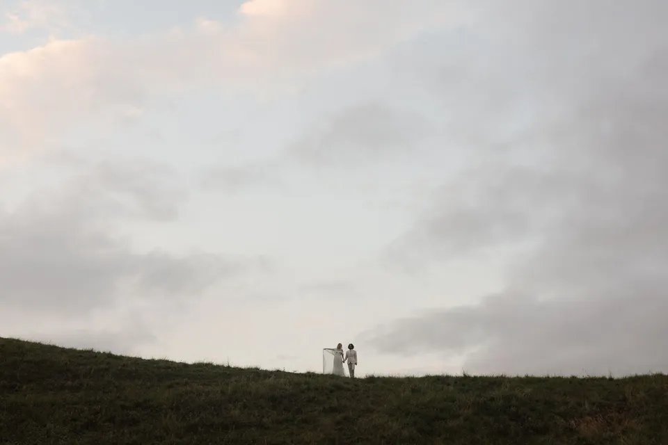 Bridget and James standing together on the hill as warm light reaches the clouds. At Cudgera Creek in northern New South Wales, for an Elopement Collective elopement. Photograph by Bulb Creative.