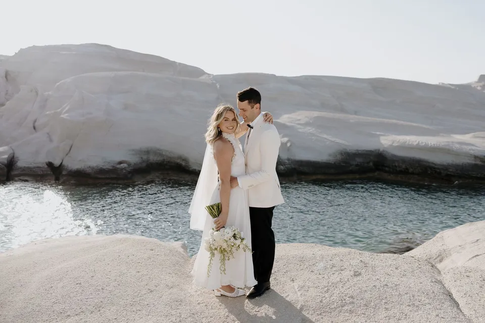 Georgina and Brad embracing beside an inlet, with white cliffs behind them and her Giasemi bouquet in her hand, at Sarakiniko on Milos in the Cyclades, Greece, during an Elopement Collective elopement. Photograph by George Bowden.