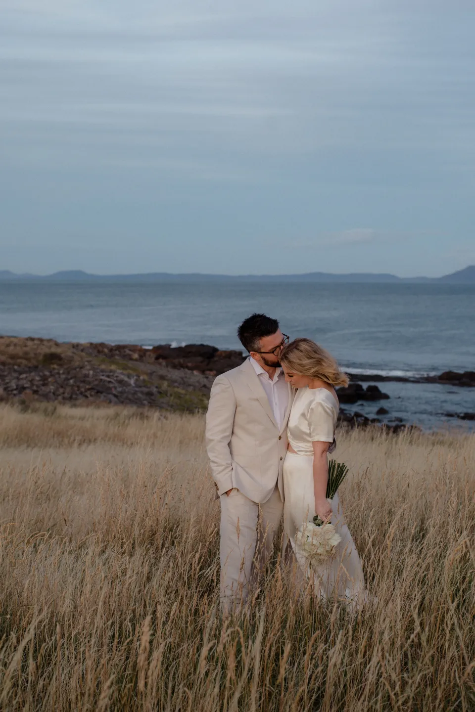 Jack leaning towards Jemmah as they stand in the coastal meadow, at Thalia Haven on the East Coast of Tasmania, Australia, for an Elopement Collective elopement. Photograph by Joey Willis.