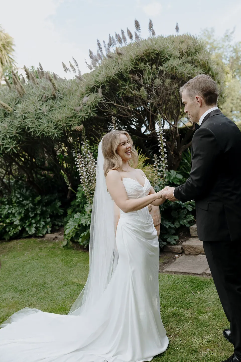 Tanesha and Andrew holding hands beneath sculpted garden trees, with her veil spread across the lawn, at Islington Hotel in South Hobart, Tasmania, Australia, for an Elopement Collective summer elopement. Photograph by George Bowden.