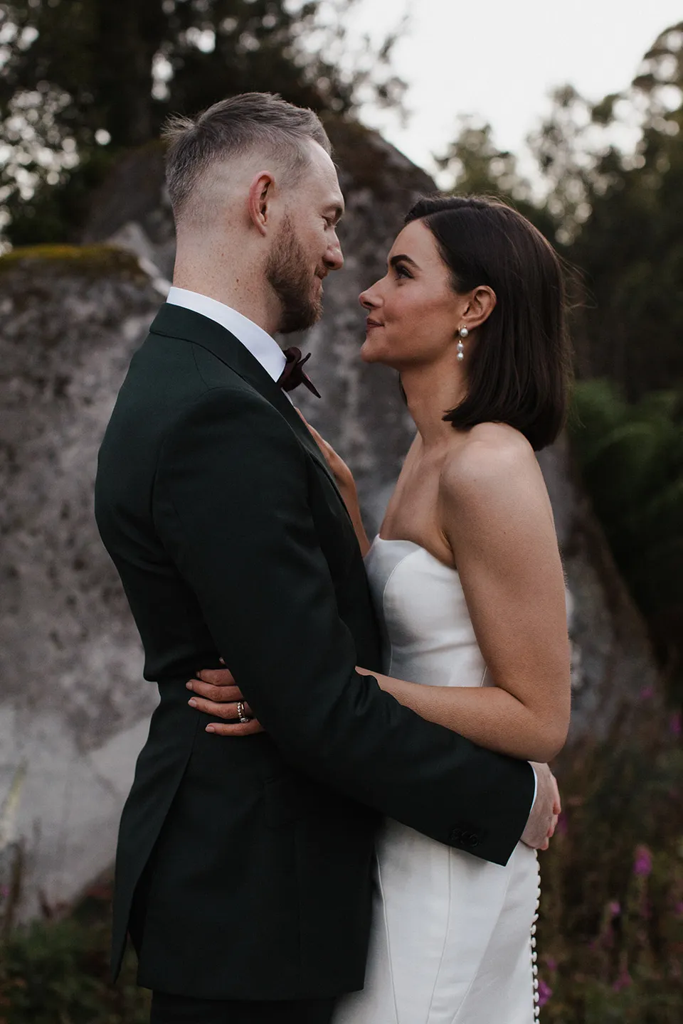 Meg and Ryan smiling at each other in a close embrace beside the woodland rocks. At The Keep on Tasmania’s east coast, for an Elopement Collective elopement. Photograph by Jenna Agius.